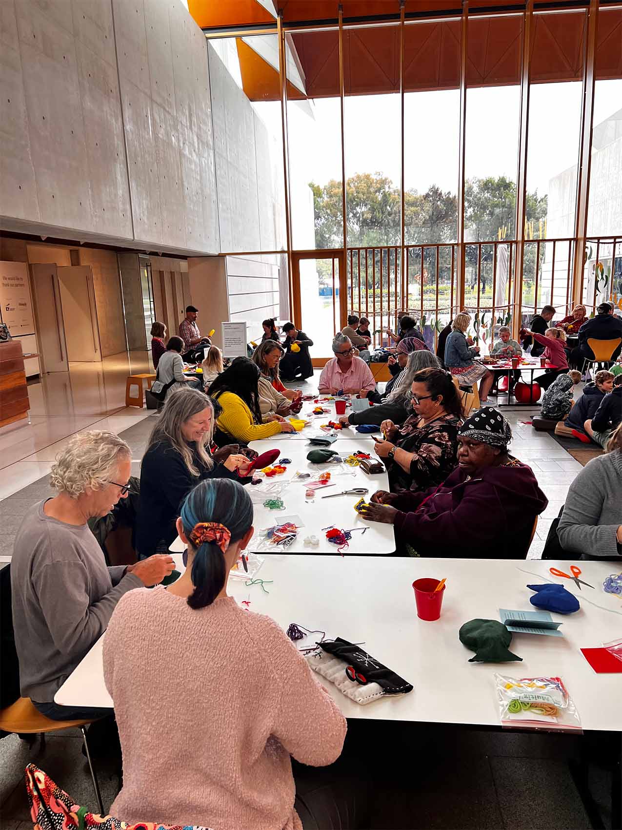 A group of people seated at tables in a bright room, engaged in crafting activities, with scissors and colorful materials on the tables.
