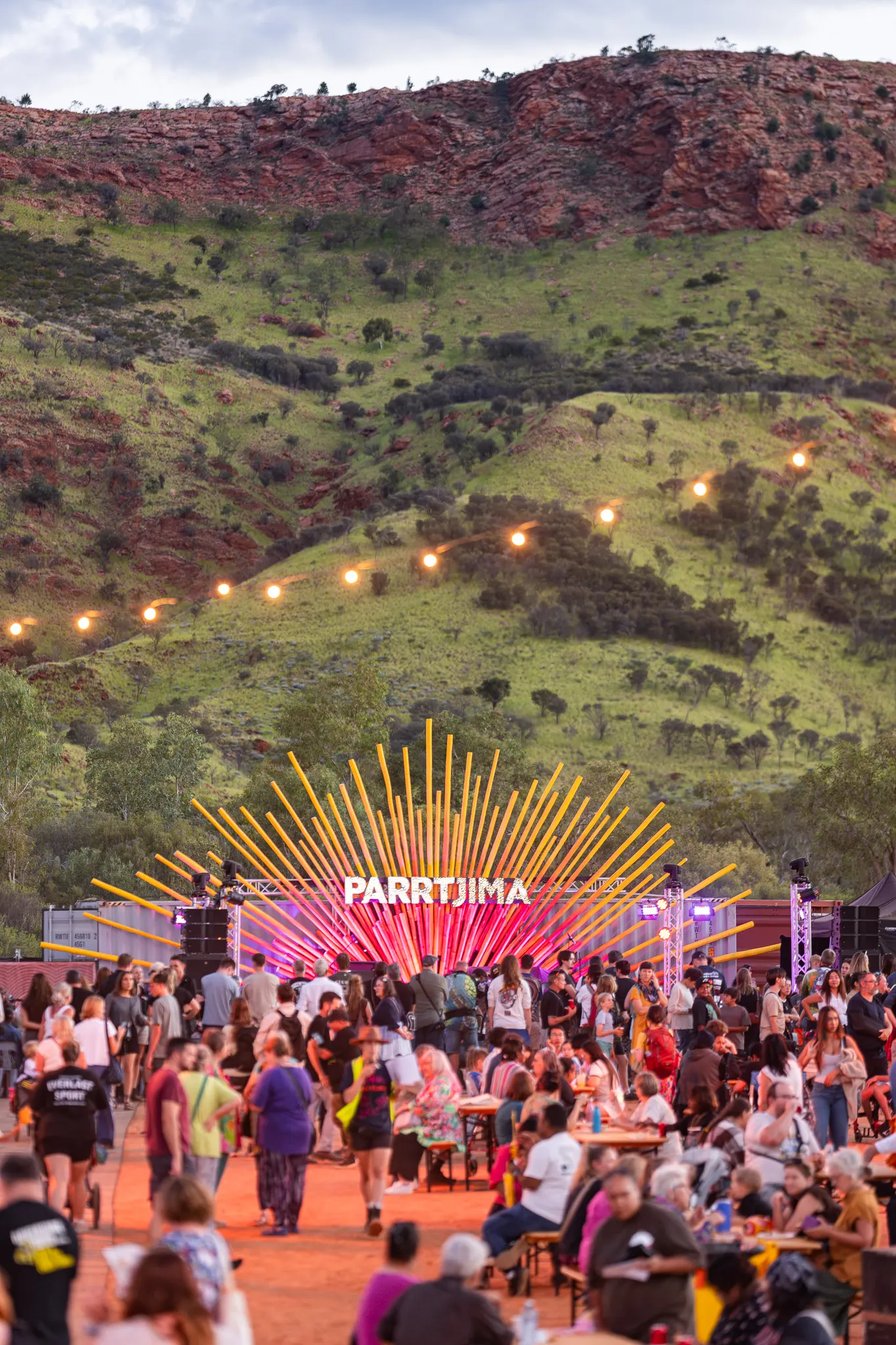 Crowd gathered at Parrtjima Festival with vibrant light installation against a backdrop of lush green hills and rocky terrain.