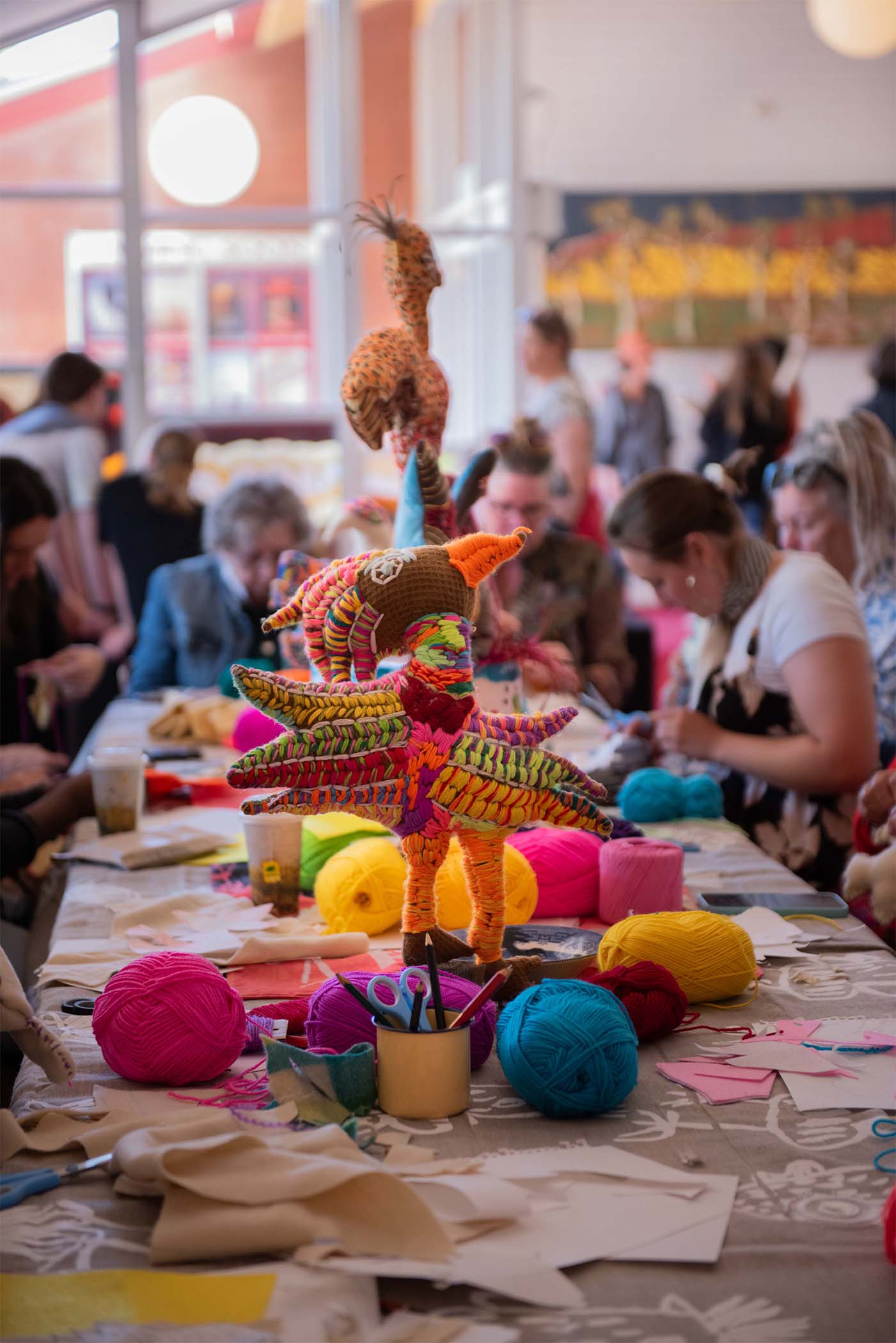 People crafting colorful wool animals at a workshop; tables filled with yarn and materials in a lively community setting.