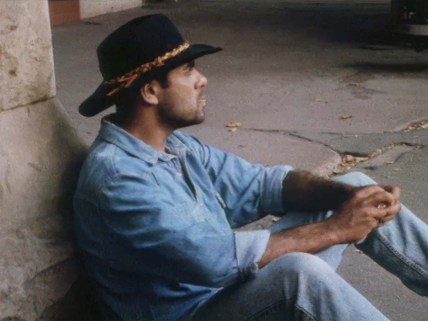 Man in denim and a black hat sits on the ground against a stone wall, looking contemplative.
