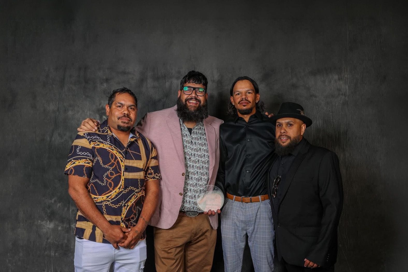 Four men stand together against a dark background, one holding an award. They are dressed in eclectic formal and casual attire.