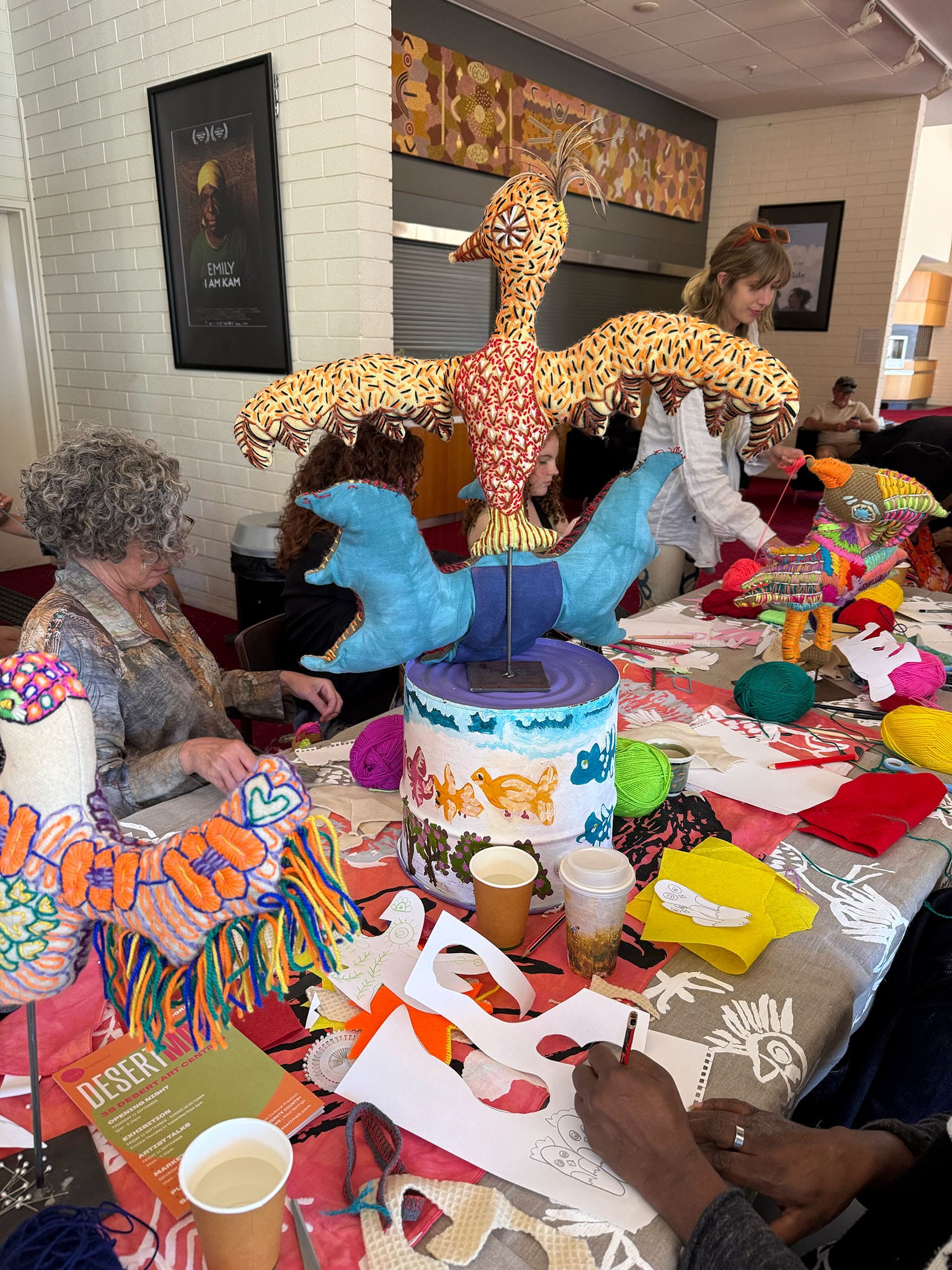 People gathered around a table creating colorful textile art with yarn, fabric, and paper. A large bird sculpture is in the center.