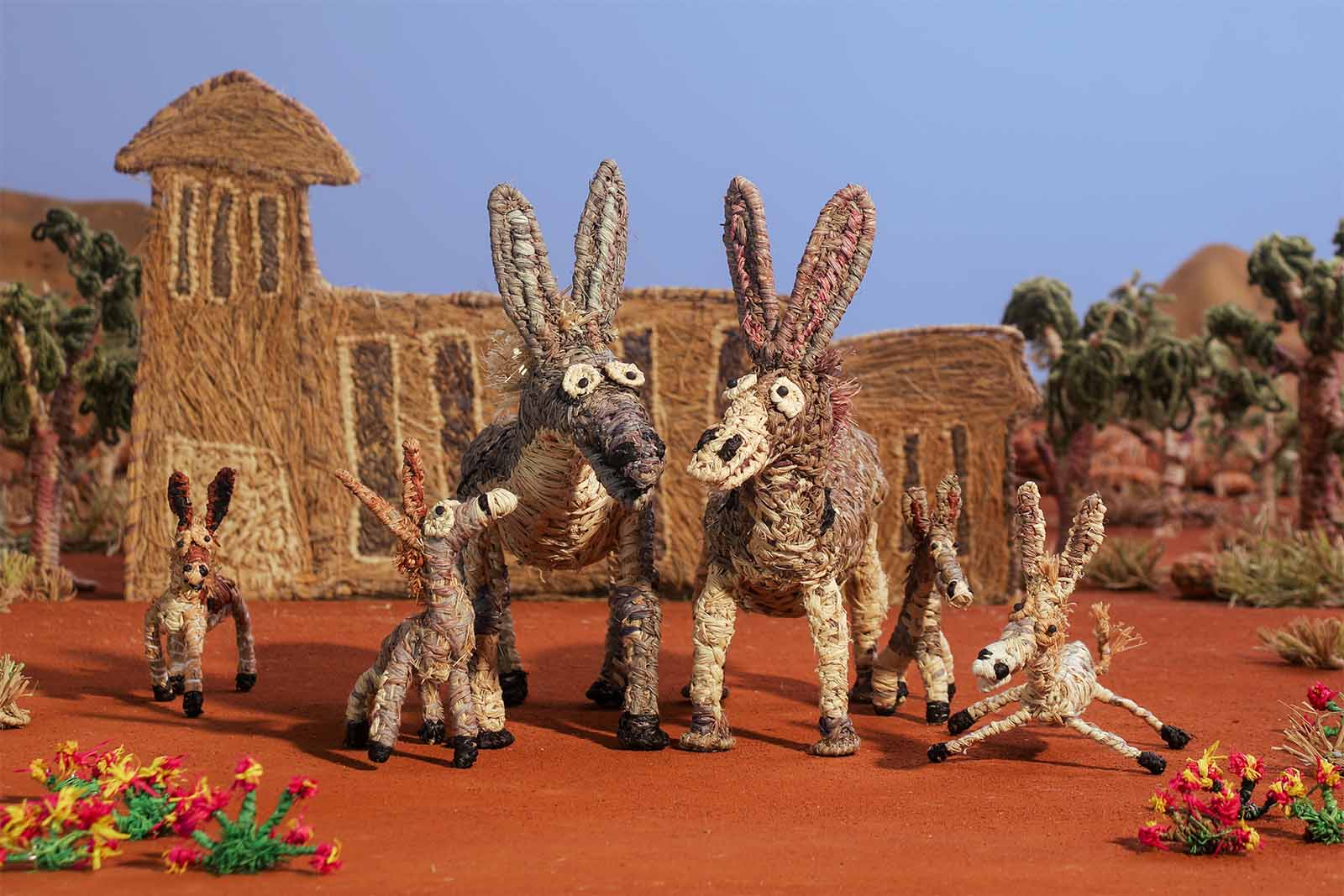 Woven donkey figures in a desert scene with a building and palm trees in the background, set on a reddish ground with colorful plants.