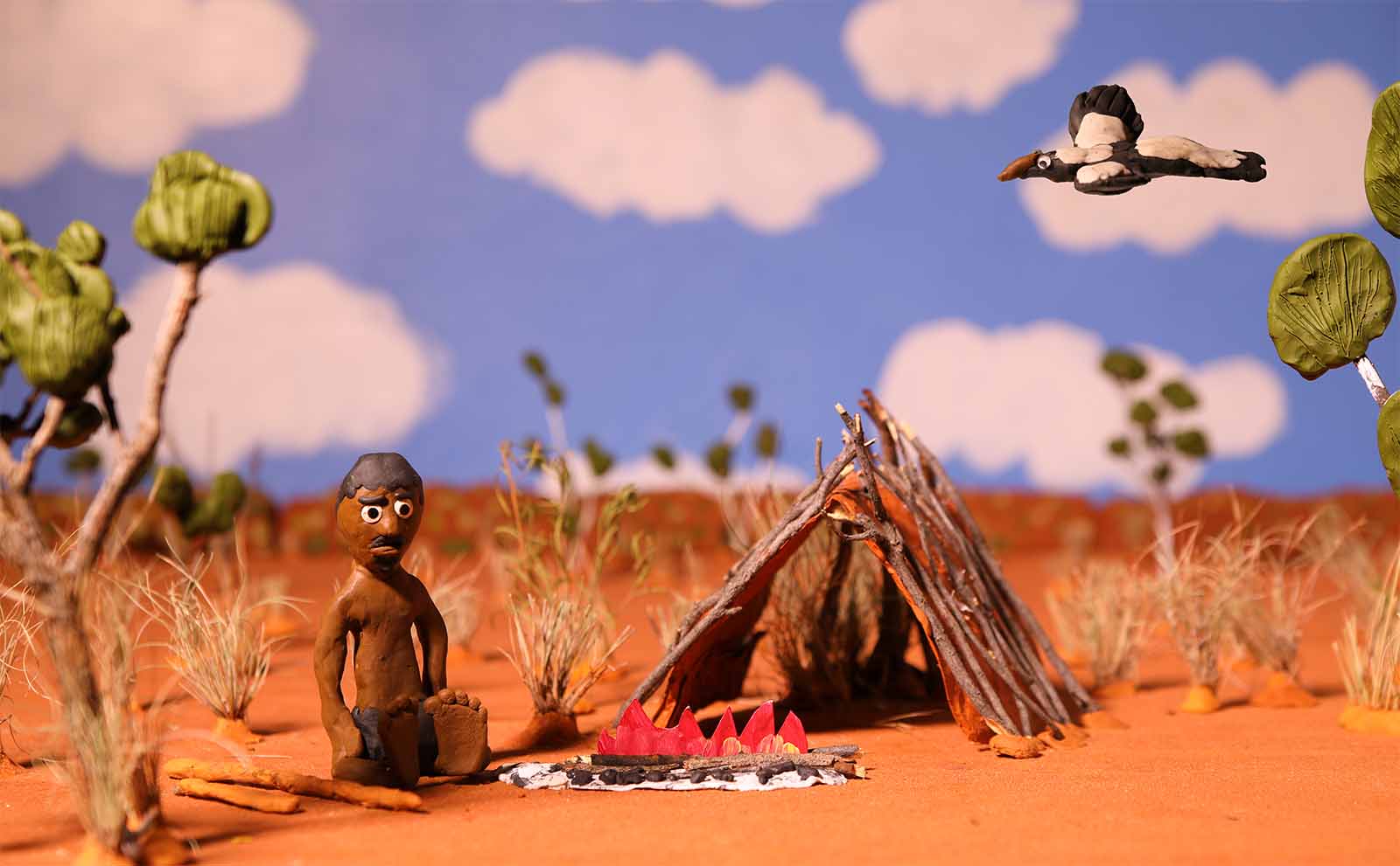 Clay figure in desert setting with stick hut and fire; bird flying above, white clouds in blue sky. Sparse vegetation surrounds.