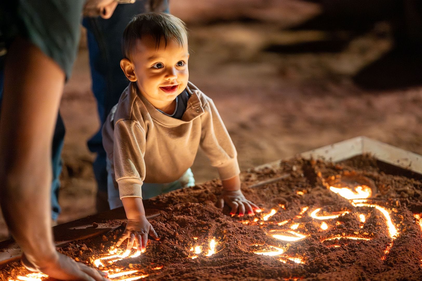 A toddler joyfully plays with illuminated sand, pressing hands into the glowing surface, surrounded by soft, warm lighting.
