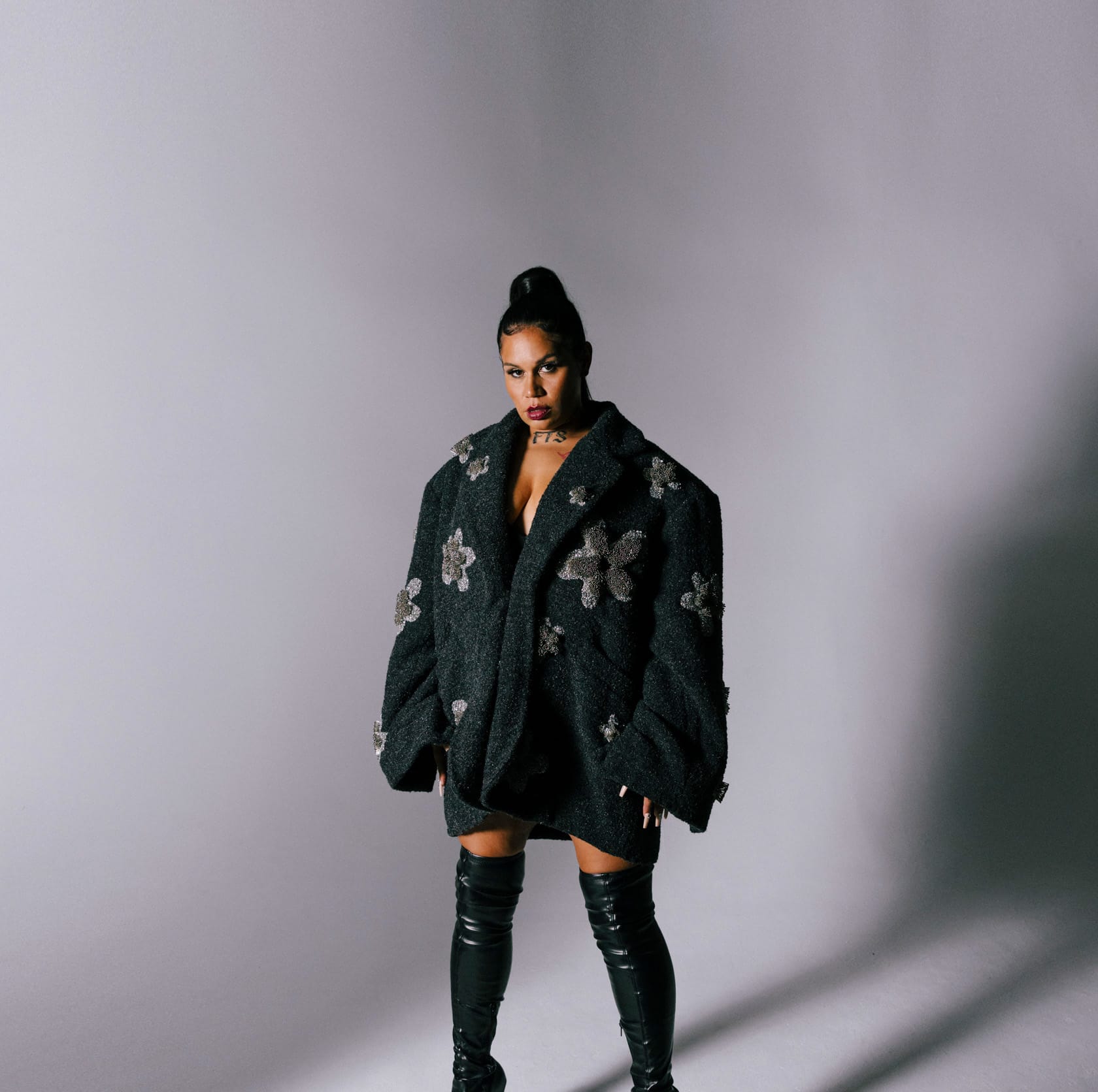 Woman in oversized floral-patterned jacket and thigh-high black boots against a plain backdrop, looking toward the camera.