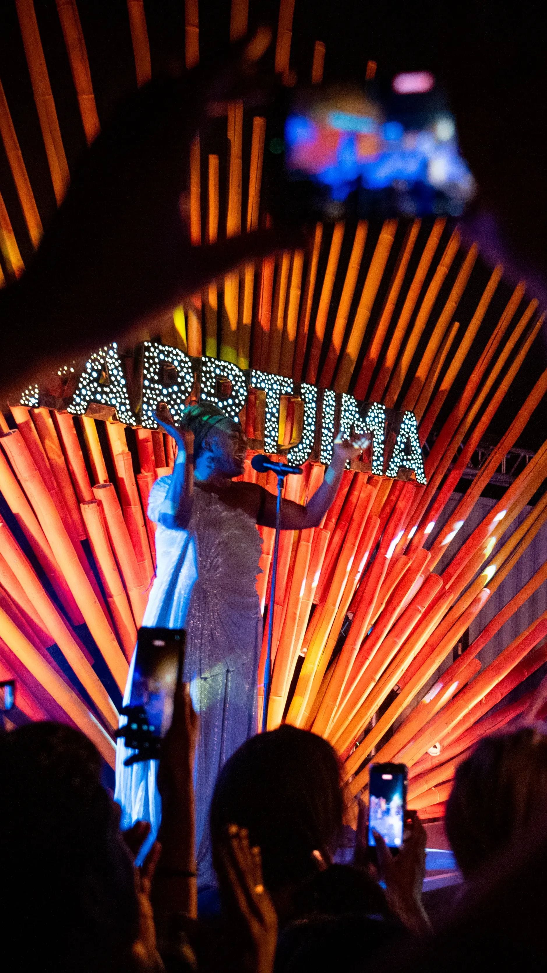 Performer in white dress sings on stage at night with orange light beams and "ARTJIMA" sign, as audience records with phones.