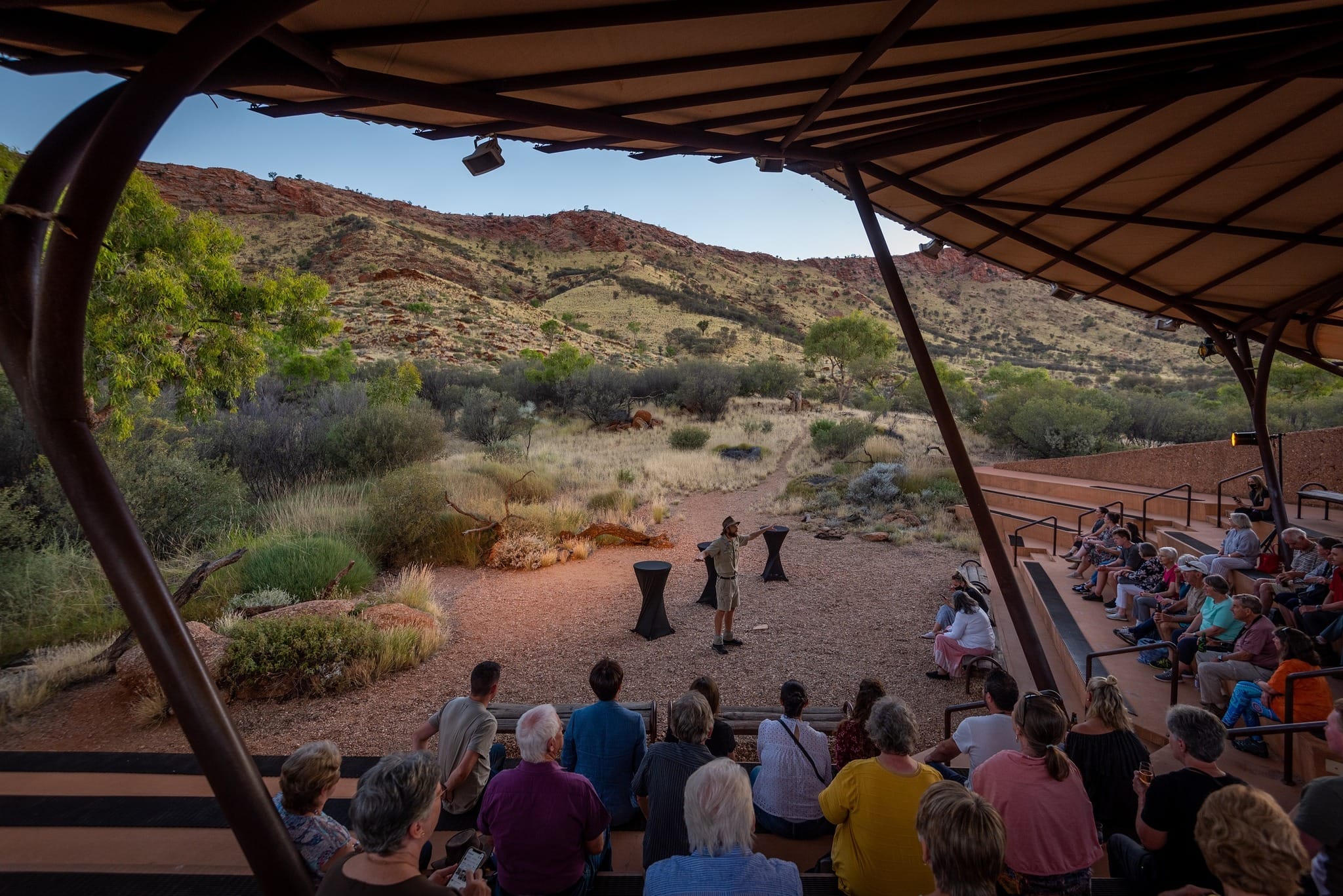 Outdoor presentation in a desert amphitheater with attendees seated, surrounded by rocky hills and sparse vegetation under a clear sky.