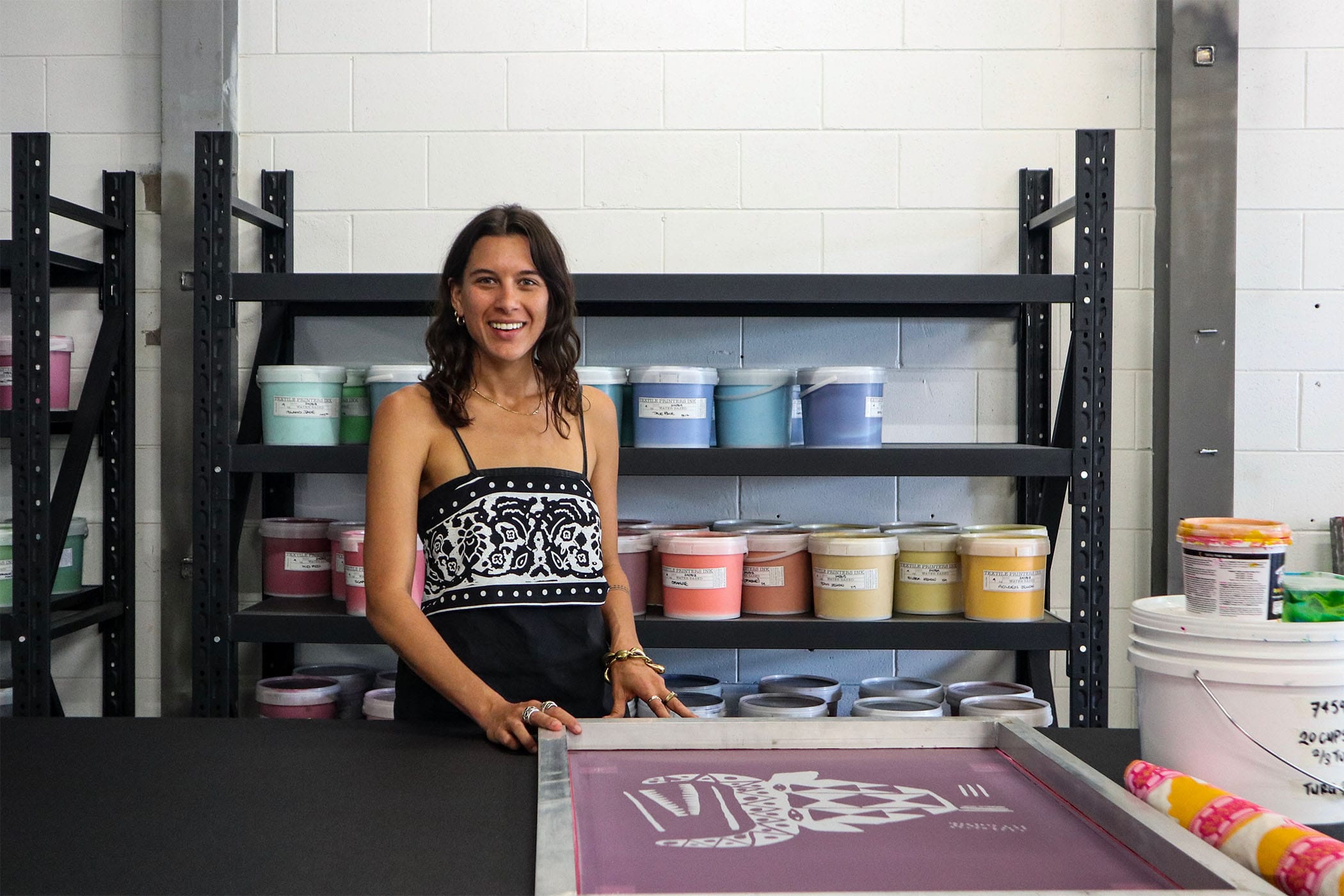 A woman stands in a workshop, smiling, with a silk screen. Shelves with colorful paint containers are in the background.