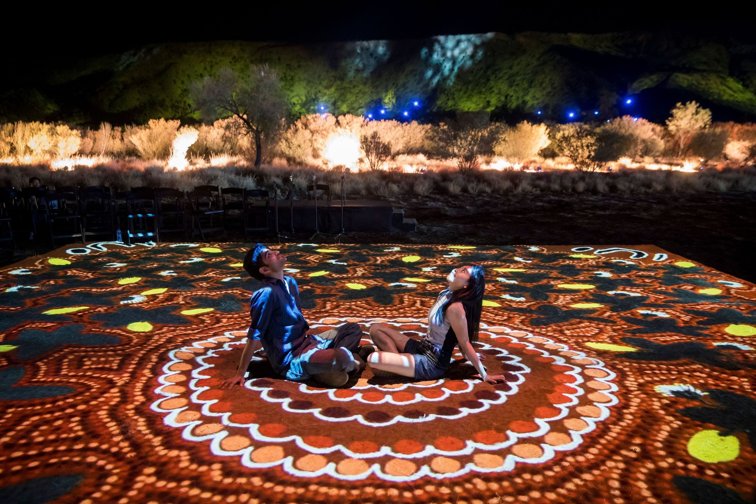 Two people sit on a decorative, illuminated ground projection with a vibrant, circular pattern in a nighttime outdoor setting.