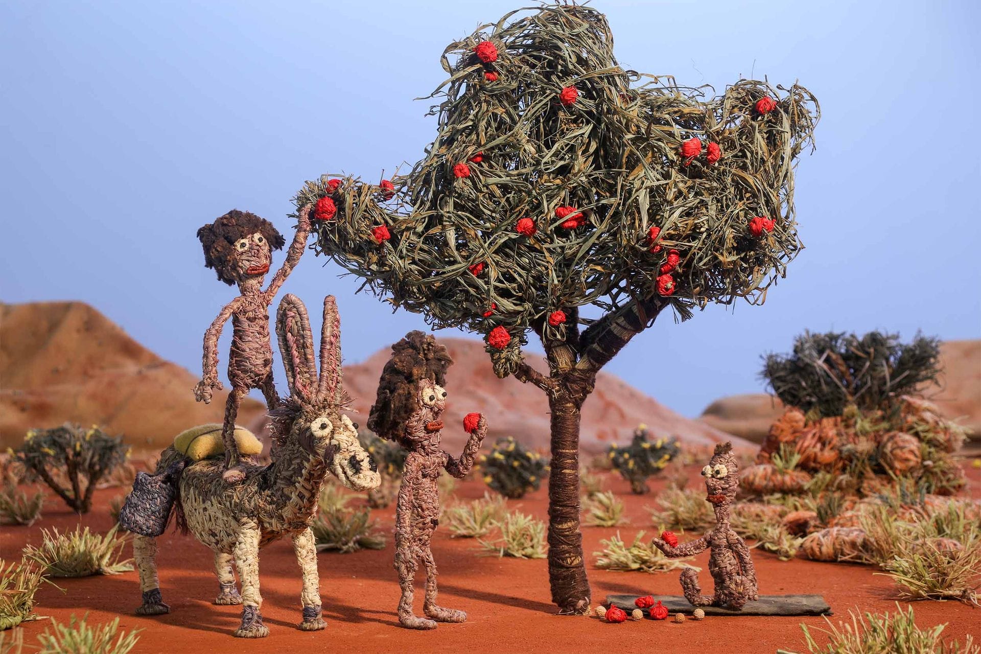 Clay sculpture scene of three figures and a donkey under a tree with red fruit in a desert landscape. One figure picks fruit while another sits.