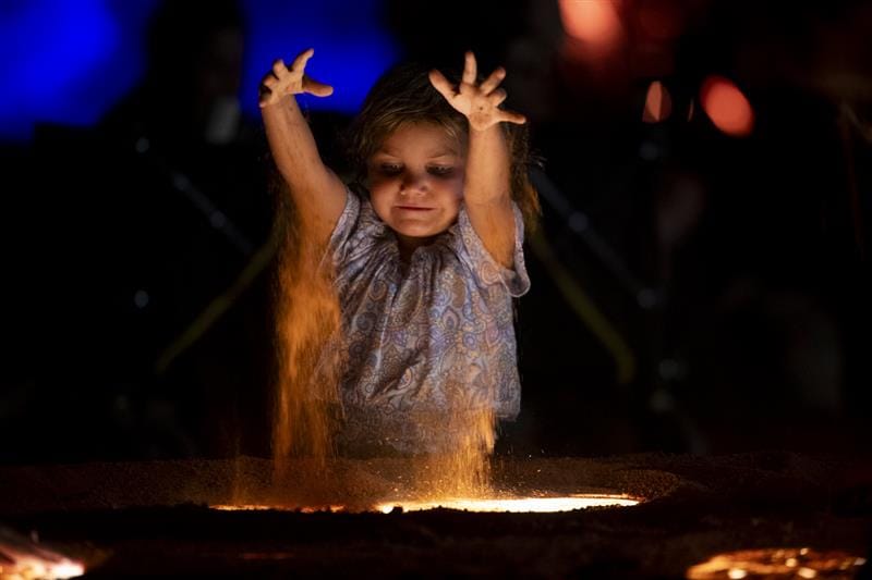 Child at night throws glowing sand into an illuminated pit.