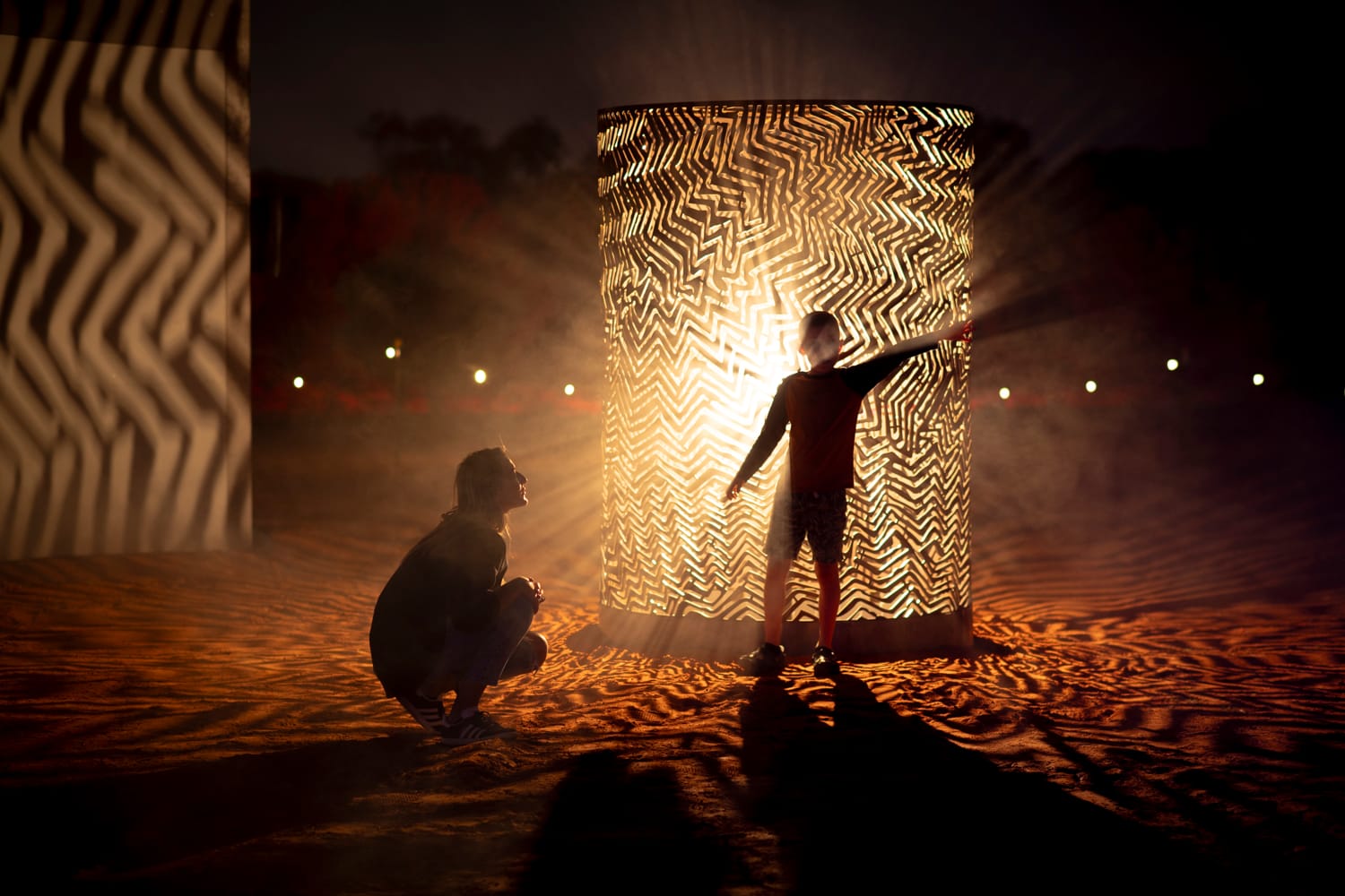 A person kneels beside a child who touches a glowing, patterned cylindrical structure, casting intricate shadows on the ground at night.