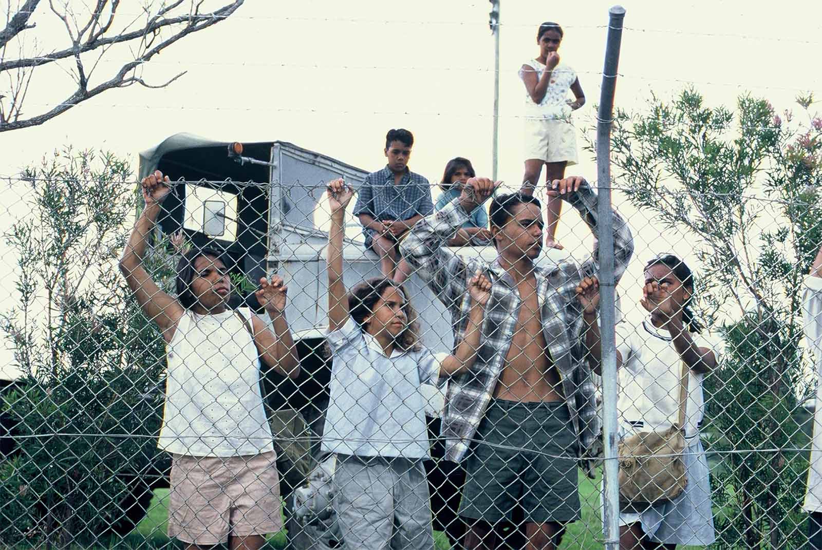 Children and teenagers stand behind a chain-link fence, peering through. An old truck and sparse foliage are visible in the background.