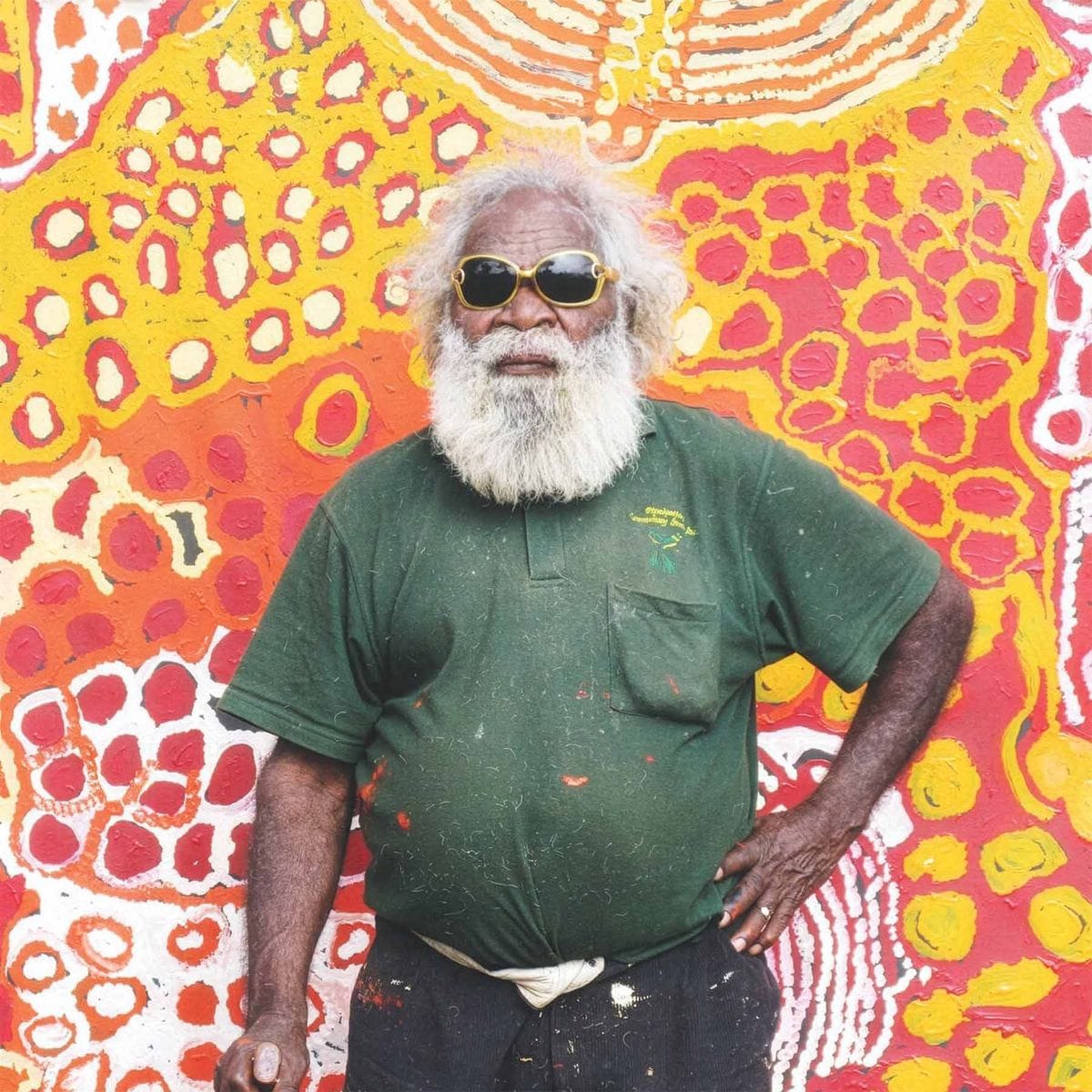 Elderly man with a white beard and sunglasses stands against a vibrant, abstract, multicolored mural wearing a dark green shirt.