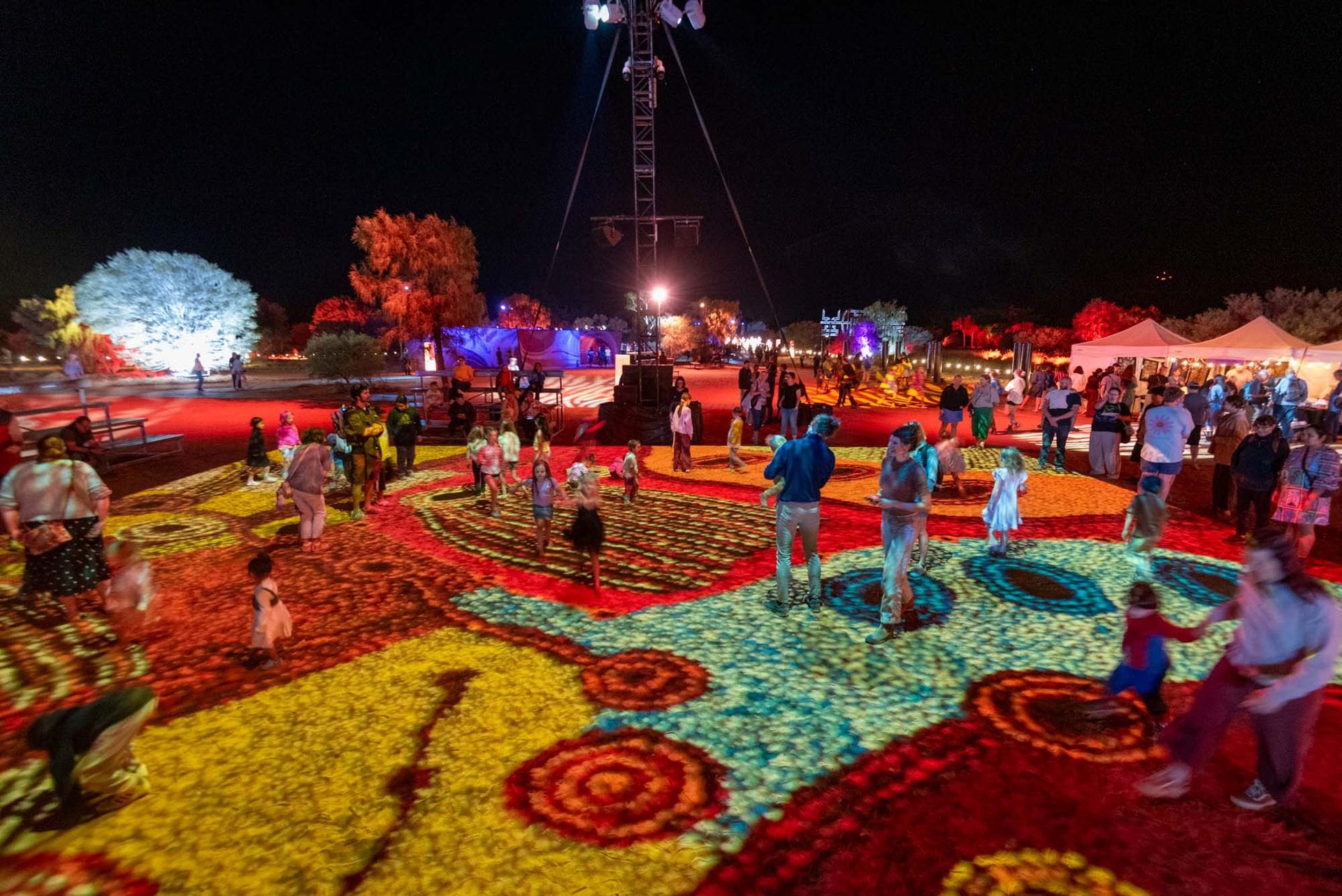 People enjoying a colorful light projection on the ground at a night festival, with trees and tents in the background.