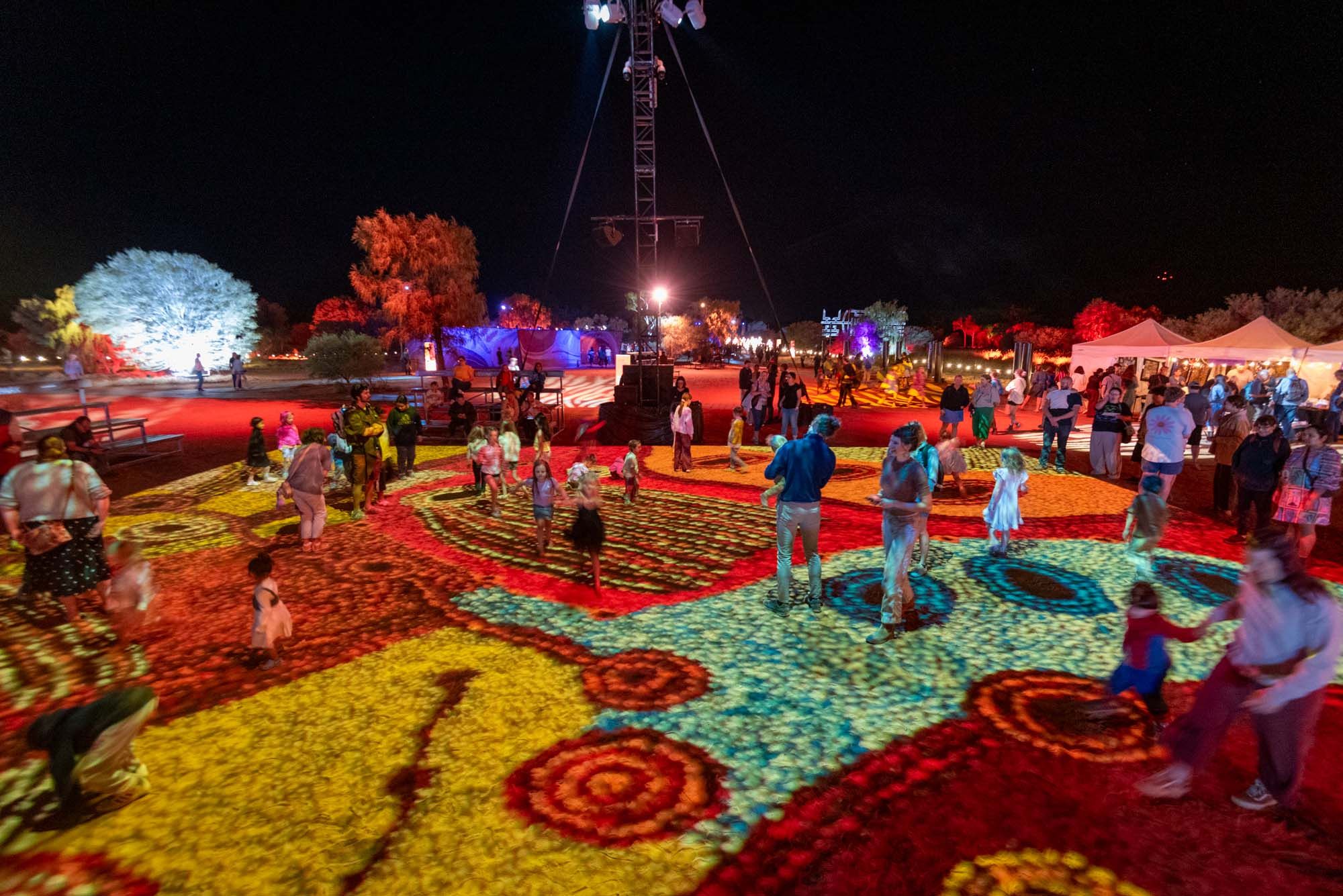 People enjoying a colorful light projection on the ground at a night festival, with trees and tents in the background.