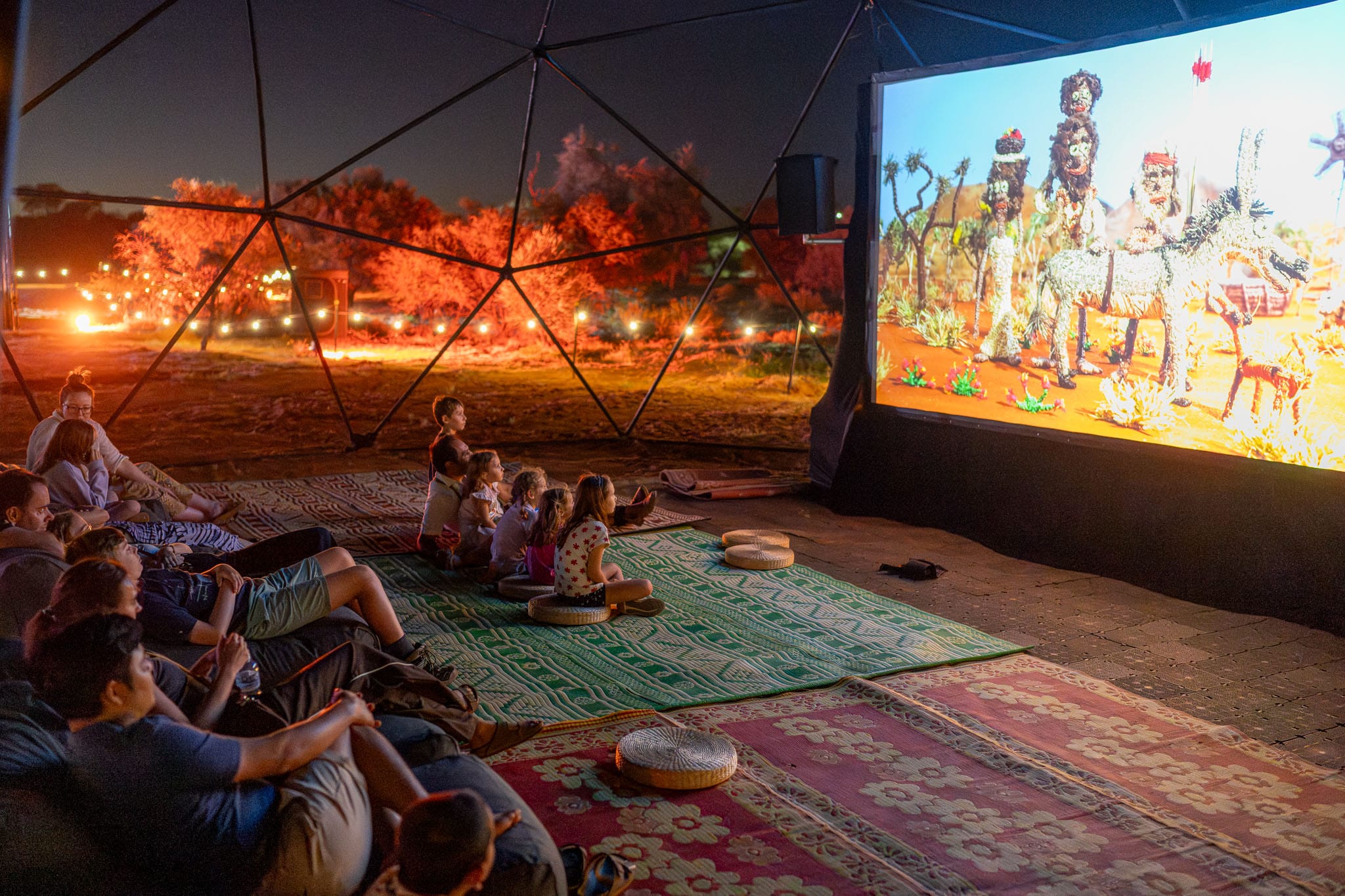 People watch an animated film on an outdoor screen in a geodesic dome, seated on colorful rugs and cushions, with trees lit in the background.