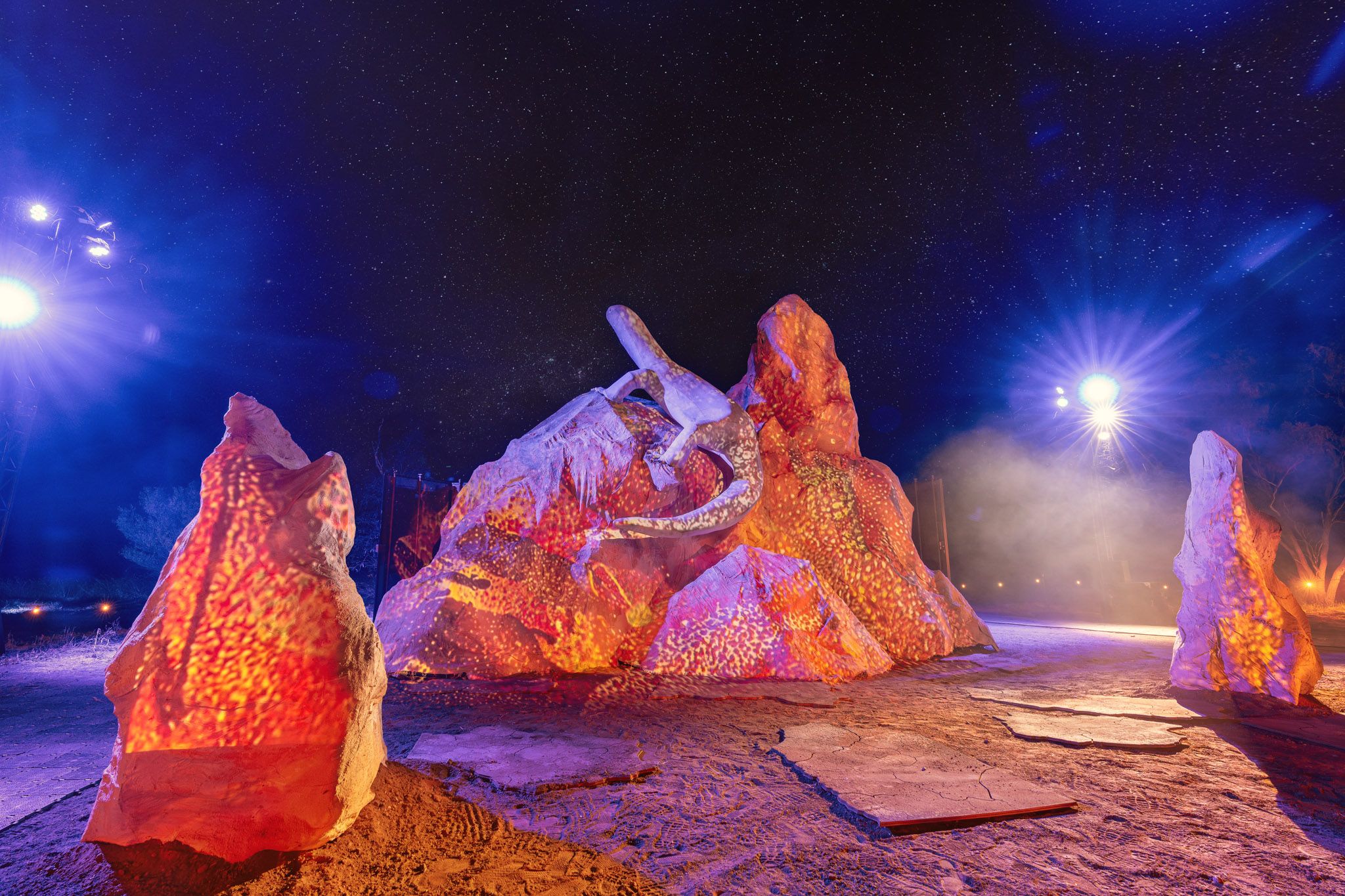Illuminated rock formations with a lizard sculpture under a starry night sky, surrounded by vibrant orange and purple lighting effects.