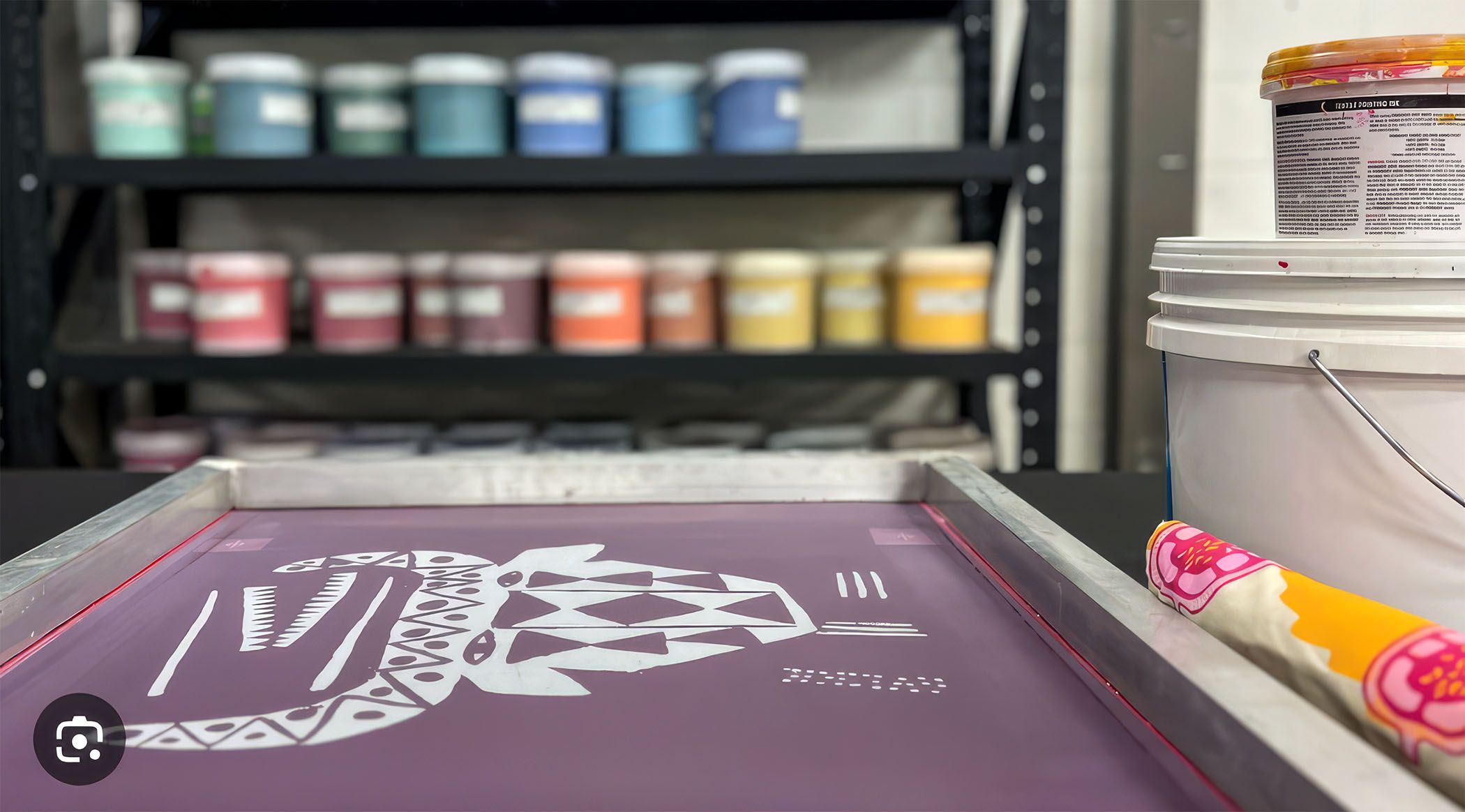 Screen printing setup with a stencil on a frame, ink tubs in the background, and a paint bucket on the right.