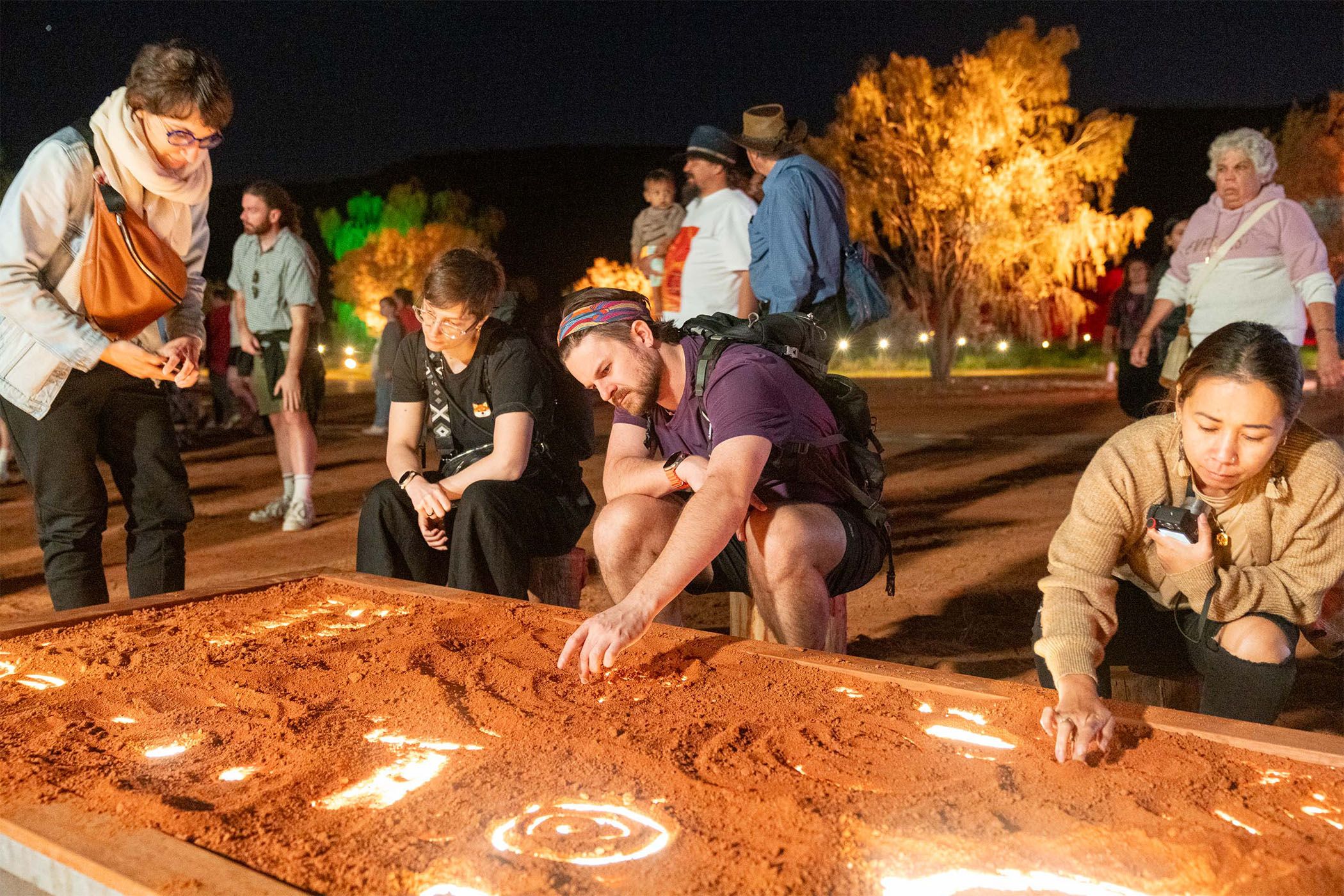 People drawing designs in sand trays under illuminated trees at a nighttime outdoor art event.
