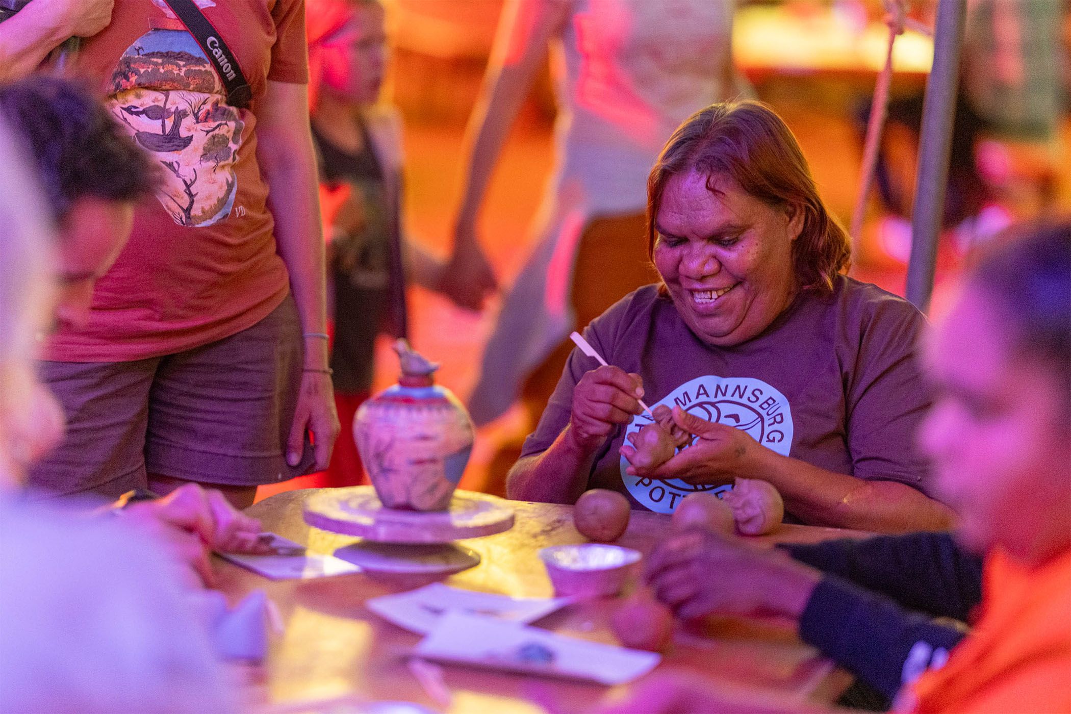 A woman smiles while demonstrating pottery-making to a seated group. The setting is warmly lit, with clay pots and tools on the table.