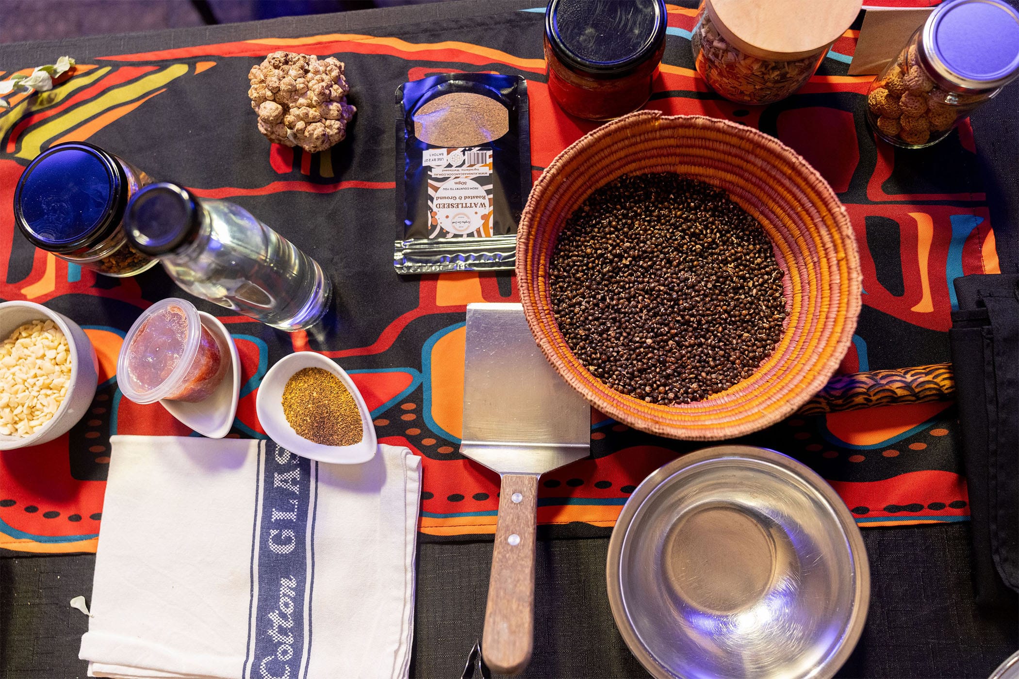 Assorted spices, grains, and ingredients on a colorful cloth, including a bowl of seeds, glass jars, and kitchen utensils.