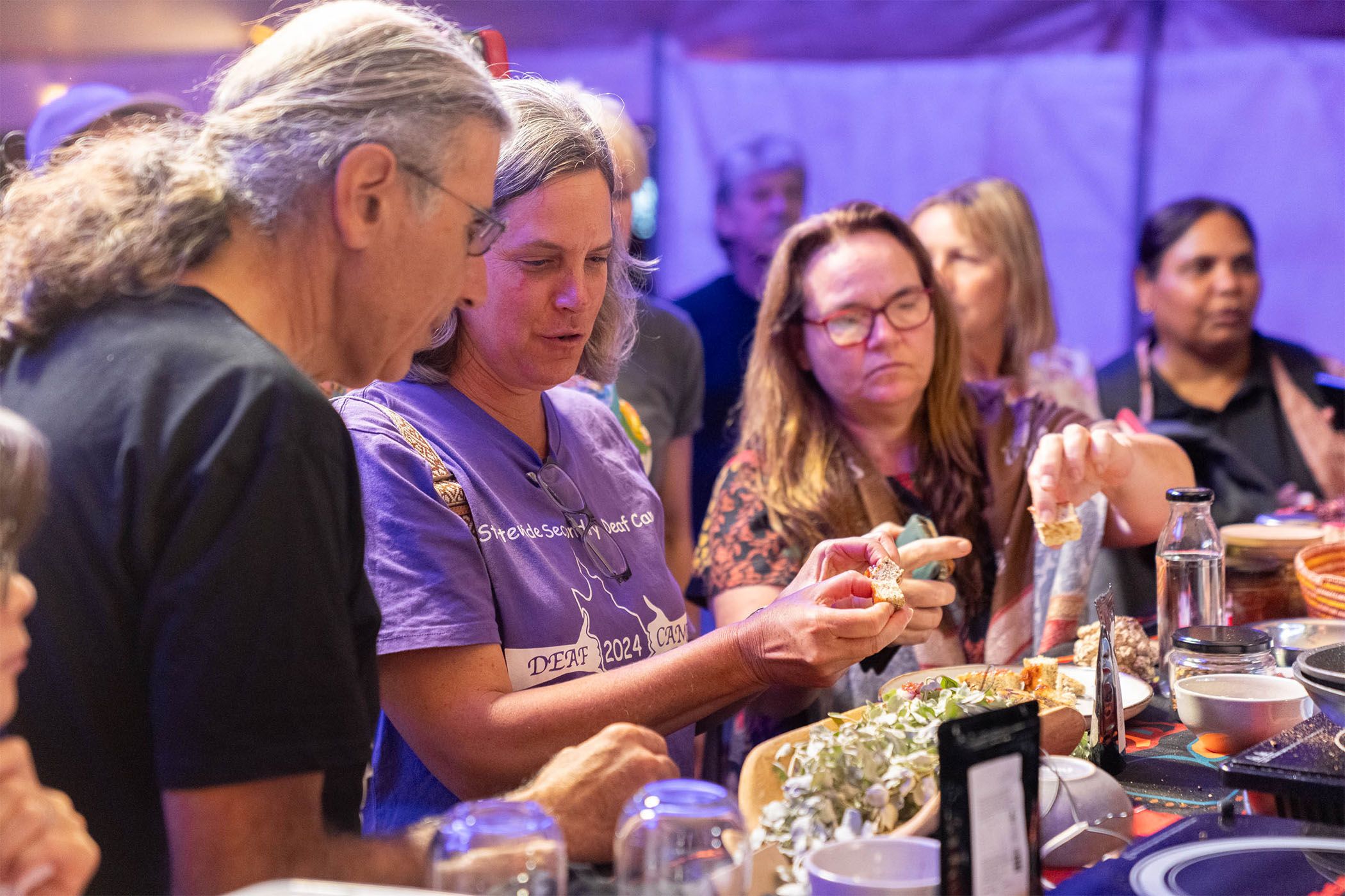 A group of people at a gathering serving themselves from a table filled with various foods, including a cheese platter and snacks.