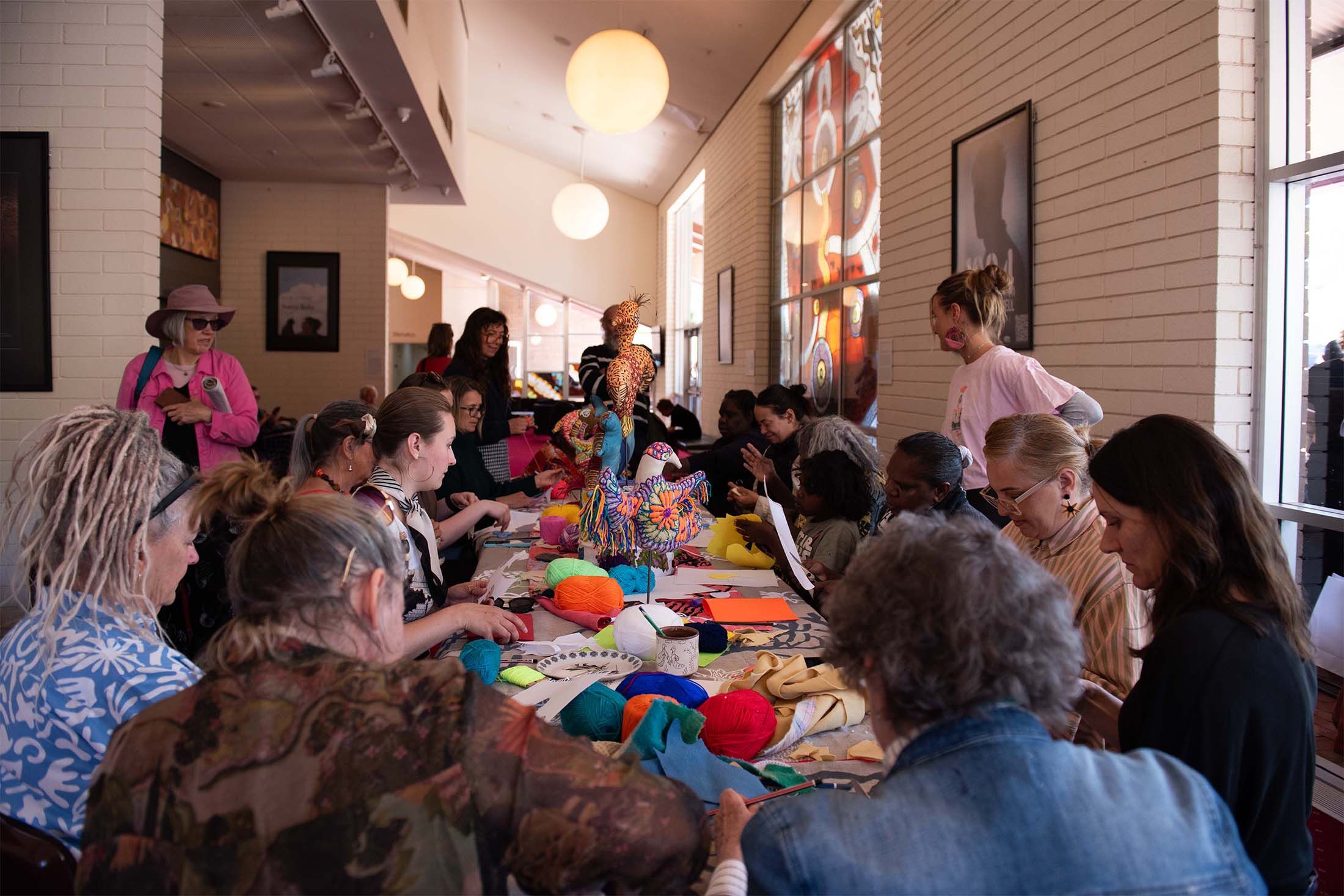A group of people gathers around a table covered with colorful yarn and fabric, engaged in crafting activities in a well-lit room.