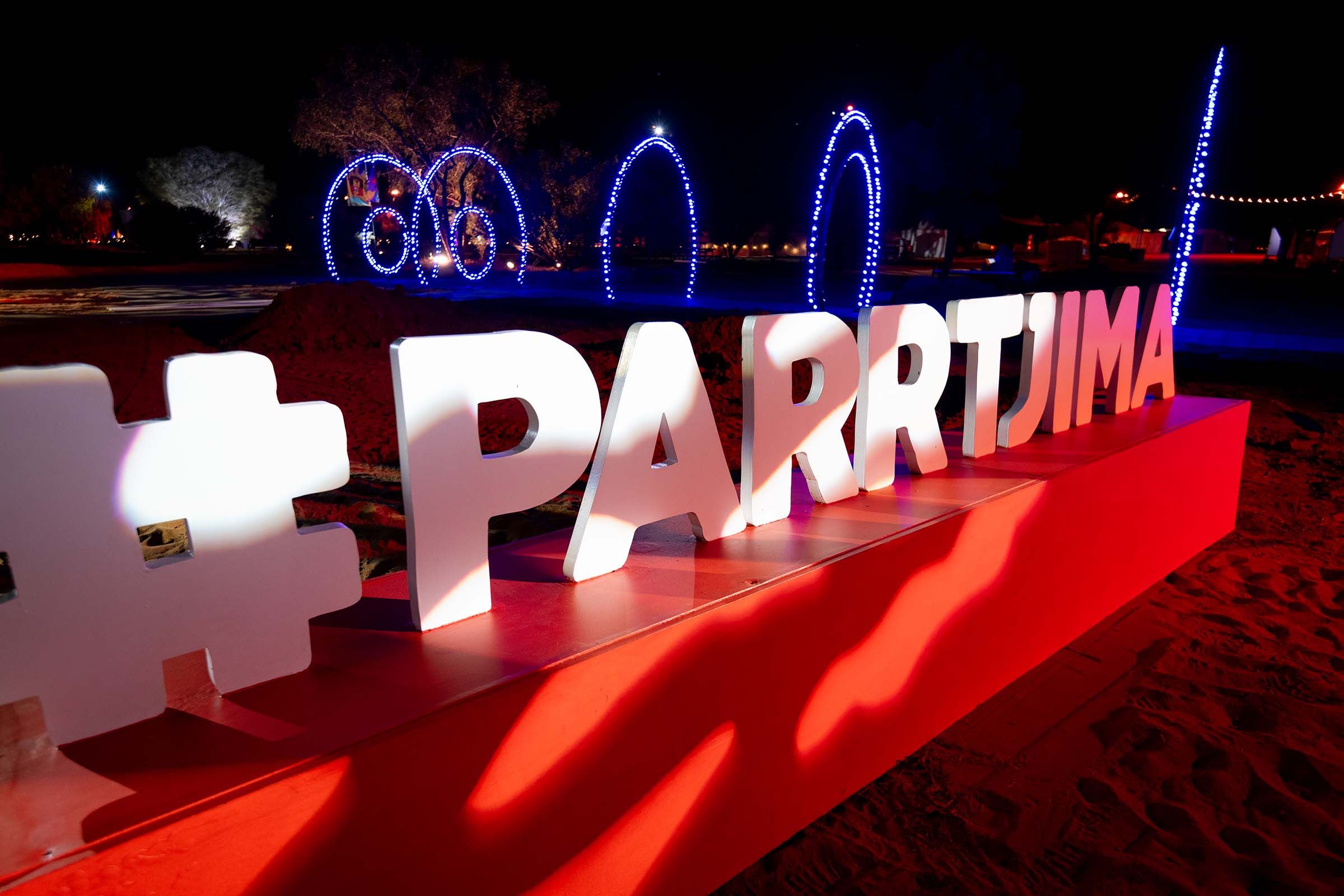Large illuminated “#PARRTJIMA” sign on a red base at night, set on sandy ground with glowing blue light installations and trees in the background.