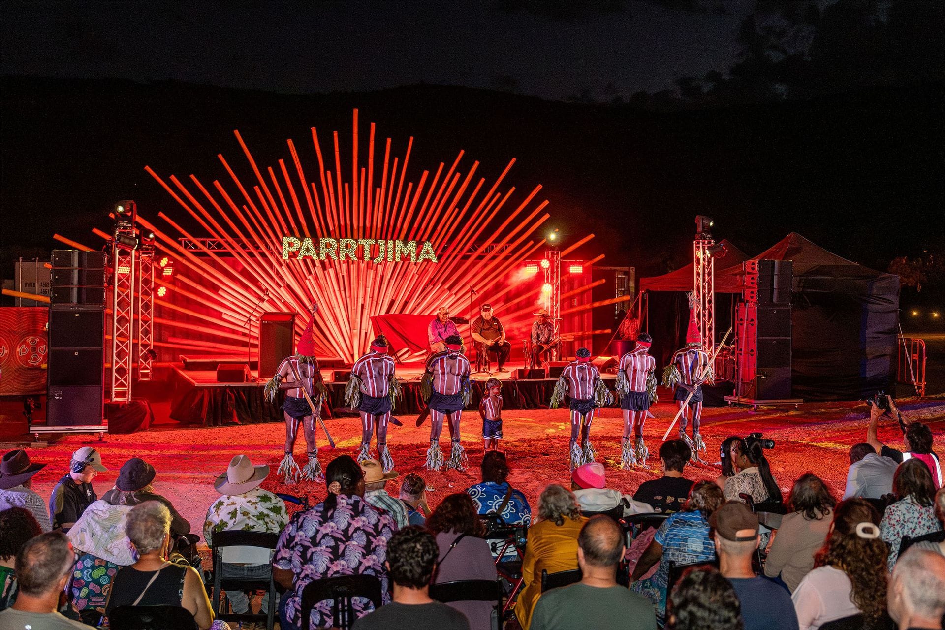 Audience watching a cultural performance on an illuminated stage with "Parrtjima" sign in red lighting at night.