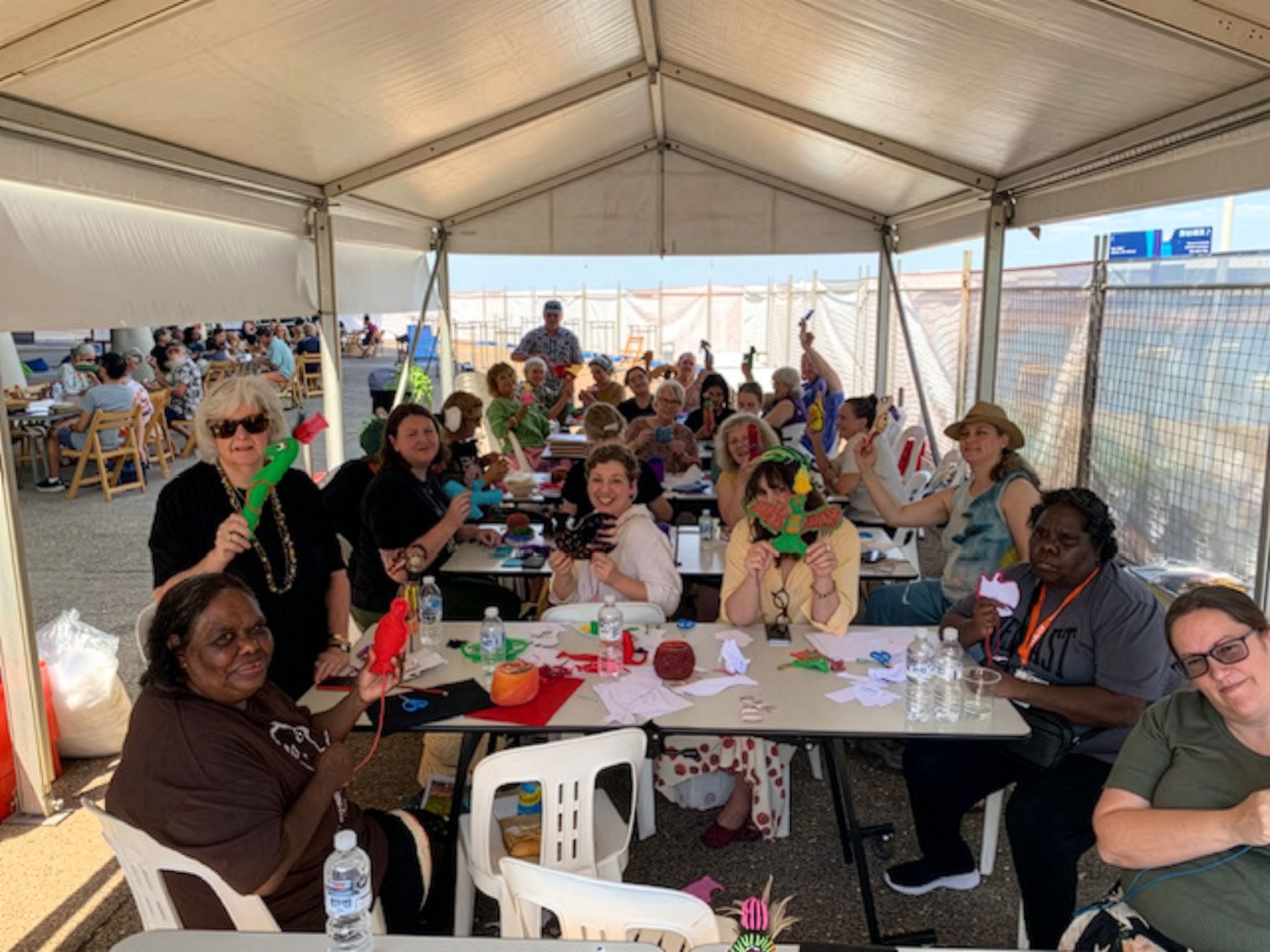 A diverse group of people sit under a tent, holding colorful craft items and smiling, suggesting a cheerful and creative workshop.