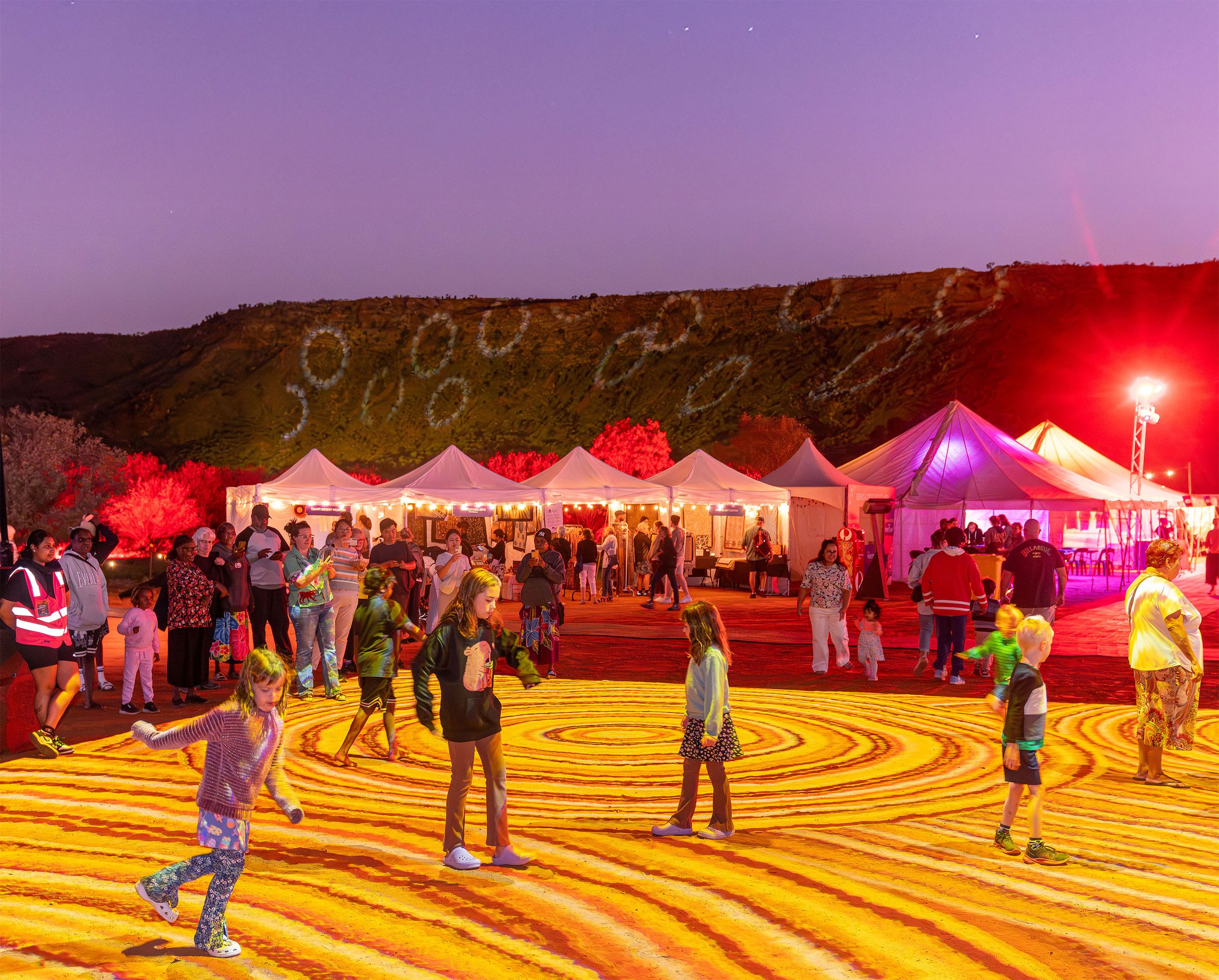 Nighttime outdoor festival with lit tents, families and children dancing on swirling yellow ground projections under red-purple lights.