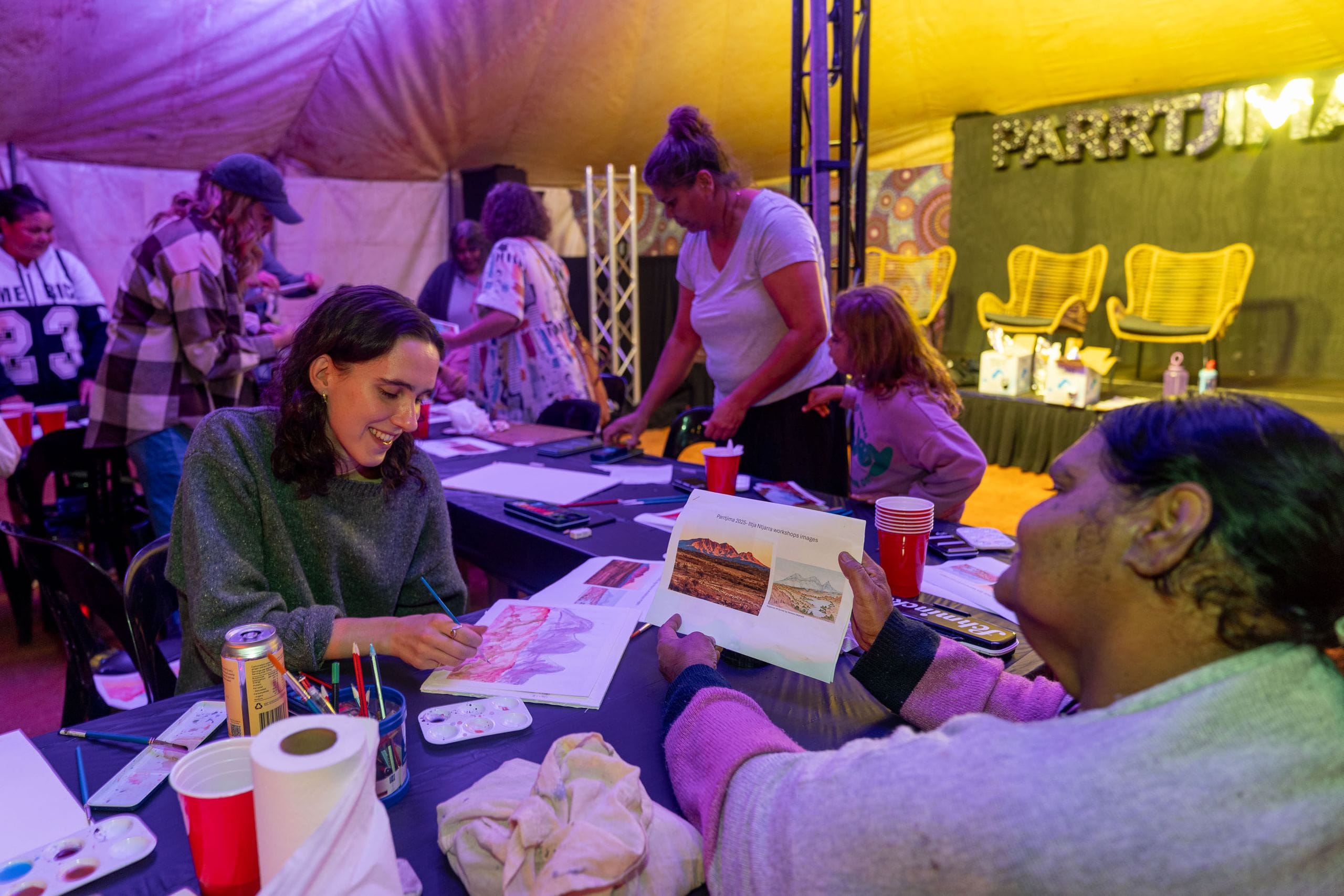 People engaged in an art workshop, painting and drawing at a table in a lively indoor setting.
