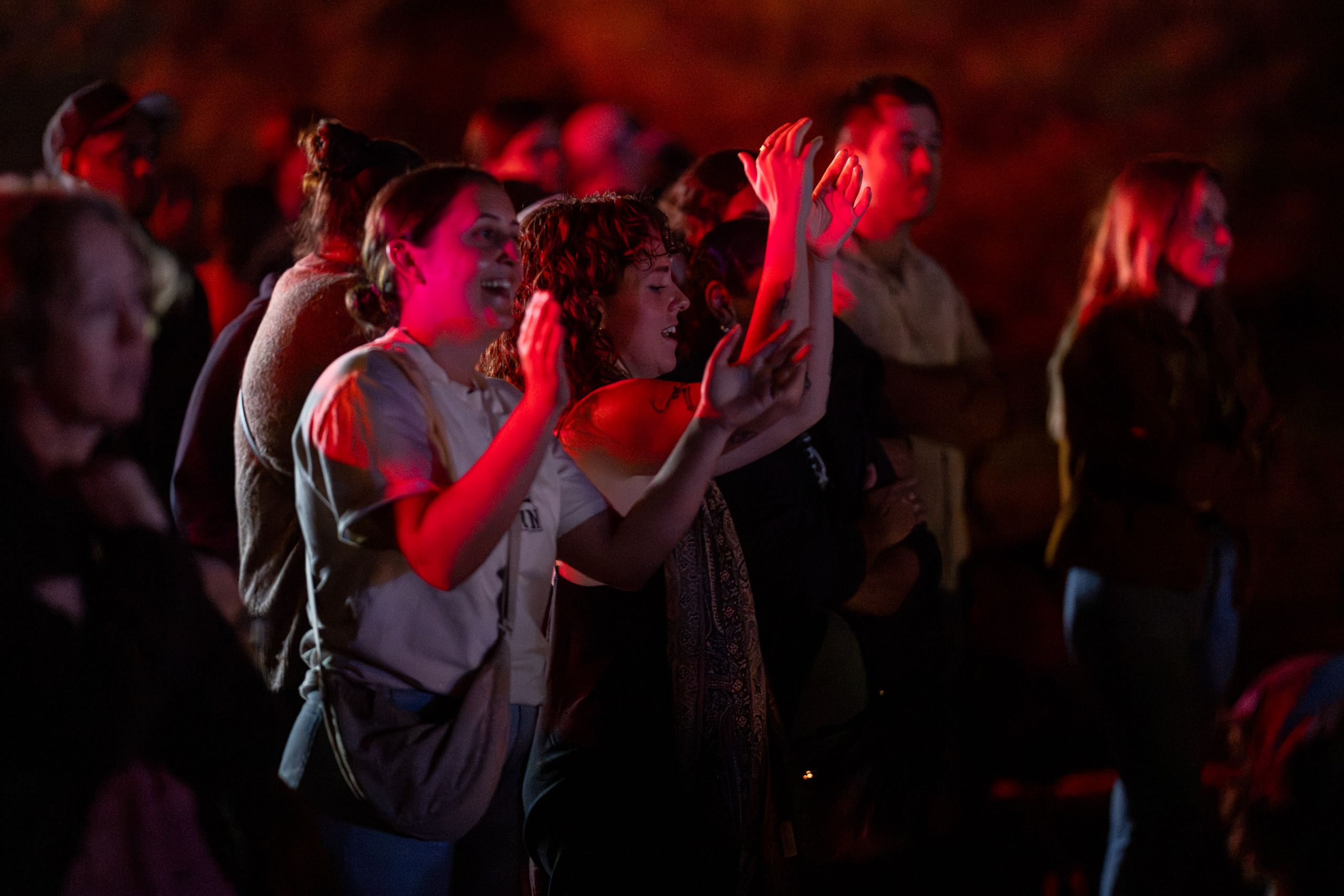 People at a nighttime concert, two audience members in front raising hands and clapping, faces lit by red stage lights.