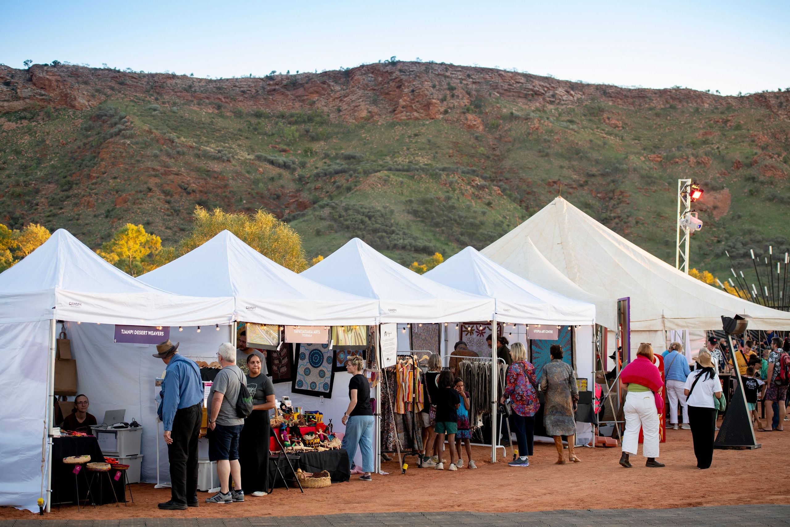 Outdoor market with people browsing stalls under white tents, set against a backdrop of green hills and a clear sky.