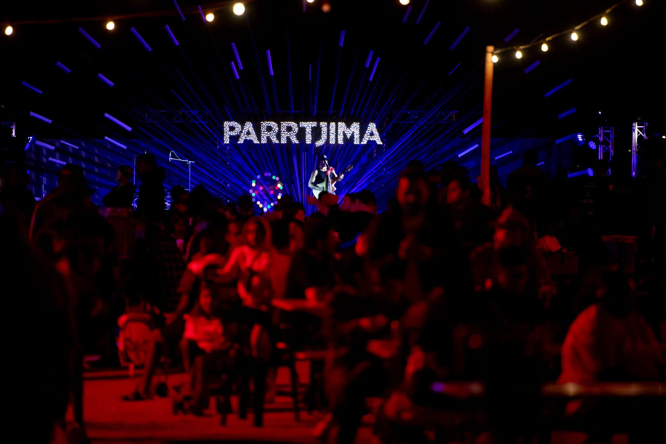 Crowd enjoying a nighttime outdoor event, with "PARRTJIMA" illuminated in the background and decorative lights overhead.