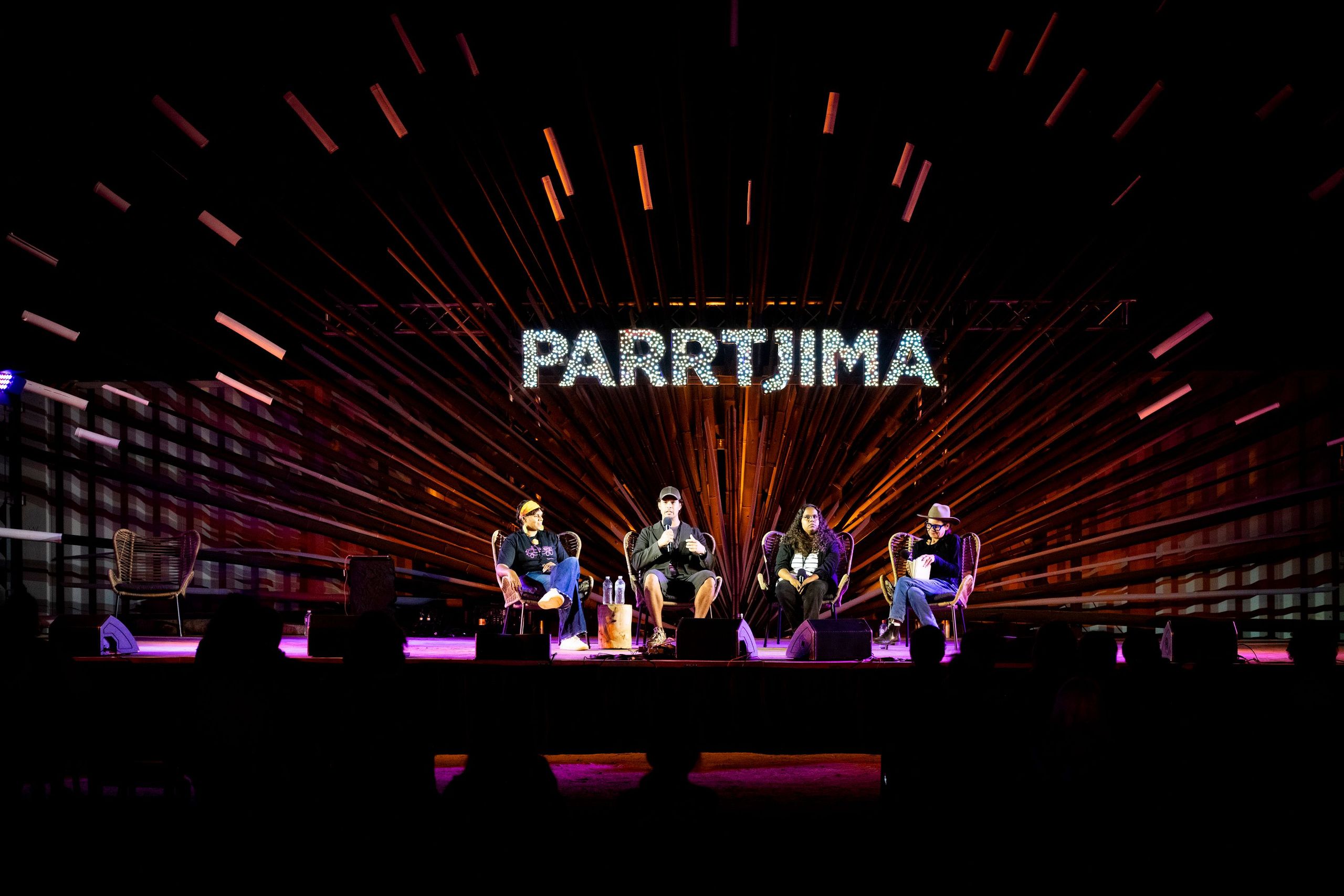 A panel of five people seated on stage with bright radial light patterns and 'PARRTJIMA' sign in the background.