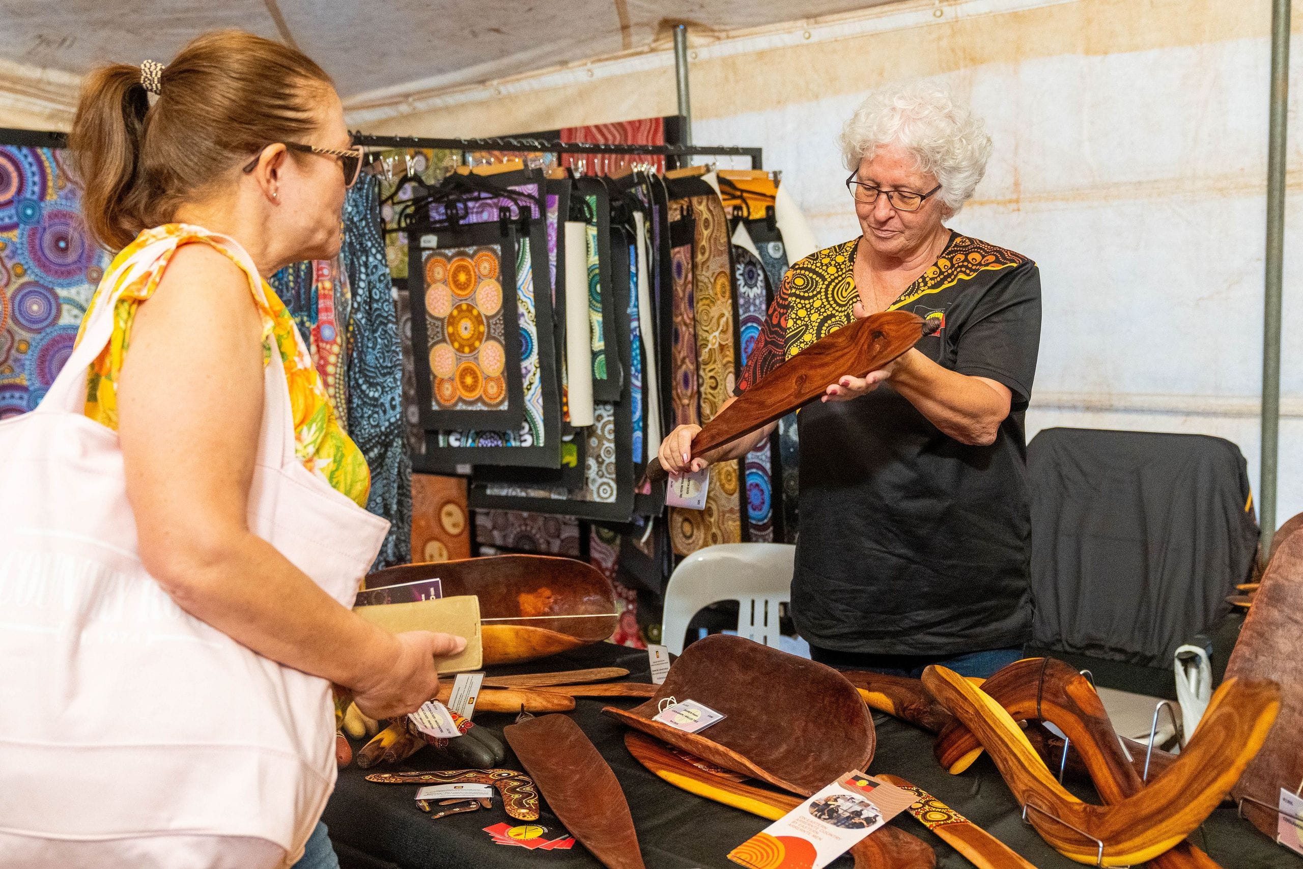 Two women in a vibrant market stall discuss handmade wooden crafts and artworks, surrounded by colorful textiles and displays.