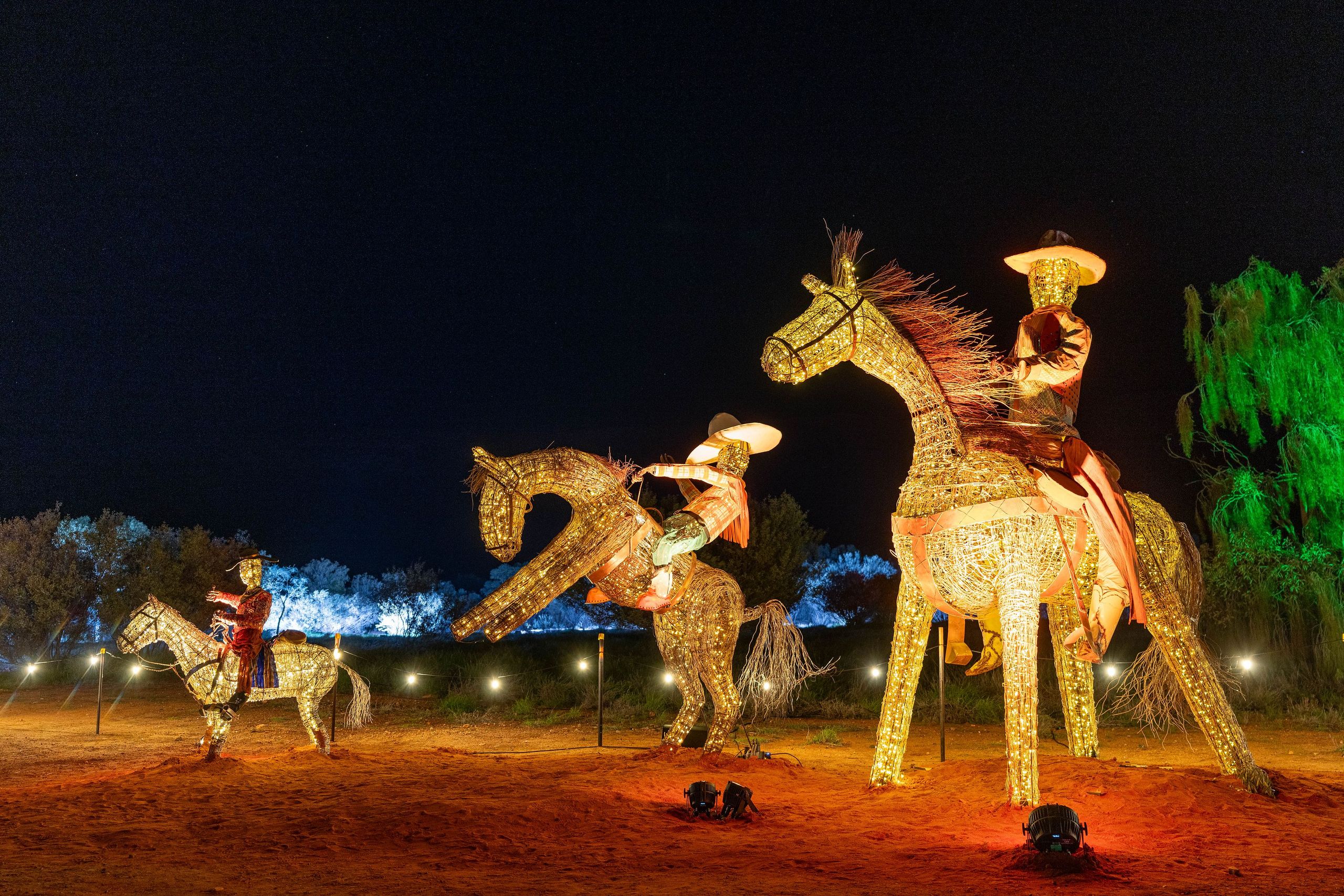 Illuminated sculptures of cowboys on horses at night, set against a dark sky with trees in the background.